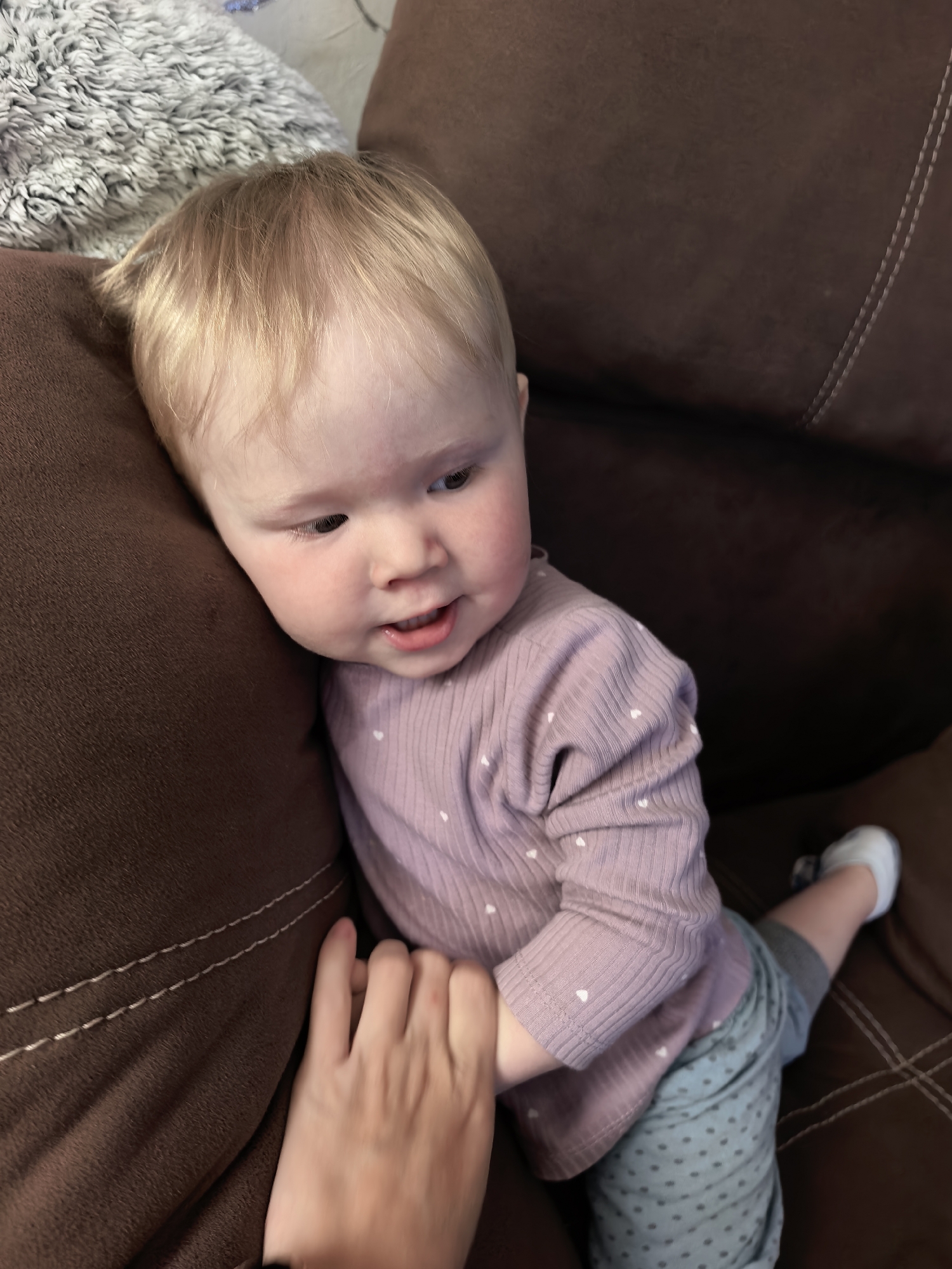 The image shows a light-haired baby or young toddler, estimated to be between 6-18 months old, seated on a brown upholstered couch or armchair. The child has blonde hair, fair skin with noticeable rosy cheeks, and dark eyes. A slight, open-mouthed smile is present as the child gazes towards the right side of the frame. The baby wears a long-sleeved, ribbed, light purple shirt patterned with small white hearts, and light blue pants with small dark polka dots. A single light grey or white sock is visible on the child's right foot. An adult's hand with fair skin is gently holding the baby's left hand against a brown cushion or armrest, which is part of or complementing the main furniture. The brown upholstery of the couch features decorative light-colored stitching in parallel lines. A portion of a light grey, textured, possibly plush or shag-like object is visible in the upper left background. The scene is indoors, appearing to be a domestic setting. The visual content of the image does not provide any indicators for its geographic location, including the city of Sterlitamak, Russia.