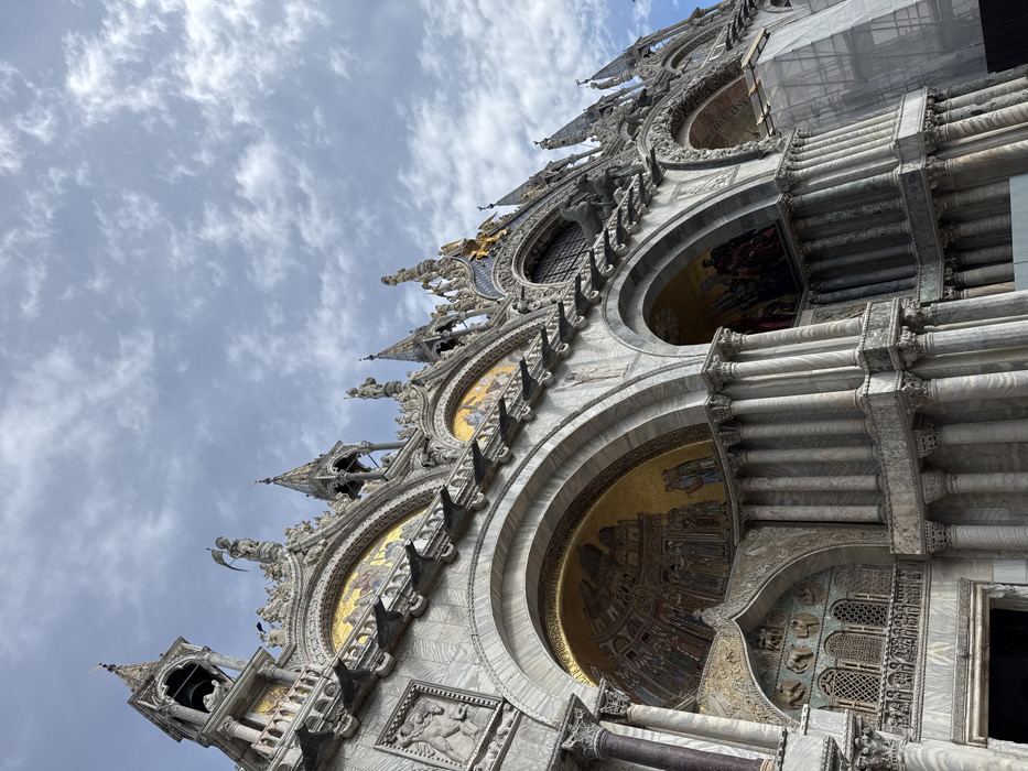 The image displays an upward-angled view of the highly ornate facade of St. Mark's Basilica, located in Piazza San Marco, Venice, Italy. The structure exhibits a complex blend of Byzantine and Gothic architectural styles, characterized by multiple arches, columns, and intricate marble cladding in shades of grey and white. Several large archways are visible, featuring golden mosaics within their tympana that depict various figures and scenes. The upper sections of the basilica are adorned with numerous statues, decorative spires, and finials, many gilded or sculpted from stone. On the far left, a distinct bell-tower-like structure with an arched opening for a bell is prominent. Above a central archway, the form of four equestrian statues, likely the Horses of Saint Mark, is discernible. The entire facade is richly textured with carvings and sculptural details. On the far right, a section of the basilica is partially obscured by grey scaffolding, indicating ongoing maintenance or restoration. The sky overhead is partly cloudy, with scattered white and grey formations. No individuals are present in the visible scene.