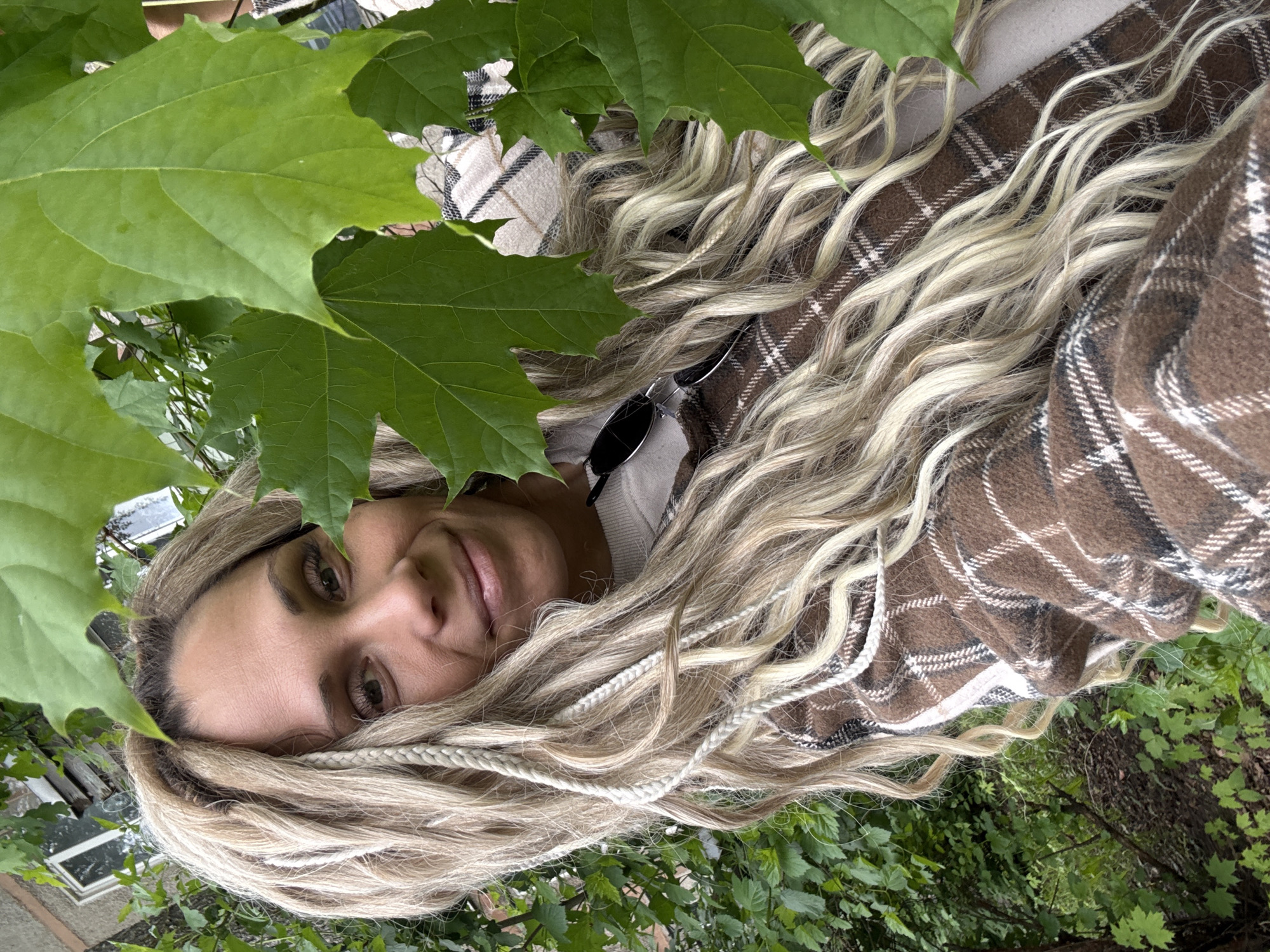 A woman with long, blonde hair intertwined with white braids is lying amongst green foliage and leaves, presumably outdoors. She is wearing a brown and white plaid garment. Her face is visible, and she appears to be looking directly at the camera. Sunglasses are tucked into her shirt. The background suggests a natural, possibly wooded, environment. Based on the prompt, the location is said to be in Rusanovo, Russia.