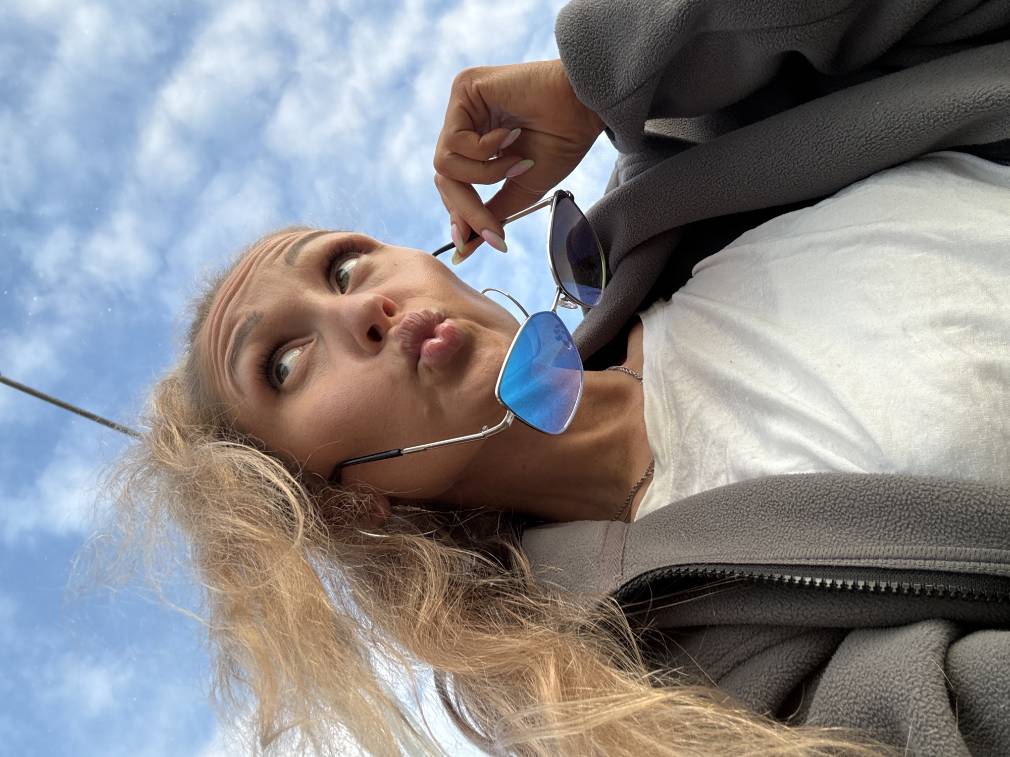 A low-angle, close-up outdoor portrait, taken in the city of Chaikovsky, Russia, features a young woman with blonde, visibly windblown hair. She is captured against a backdrop of a bright blue sky with scattered white cumulus clouds. The woman is looking upwards with her eyes, while her lips are pursed in a "kissing" or "duck face" expression. Her forehead displays slight furrowing. She is wearing a dark grey textured fleece jacket, partially unzipped, over a white t-shirt, revealing a thin silver chain necklace. In her right hand, she holds a pair of silver-framed aviator sunglasses with reflective blue lenses, with her fingers showing pastel pink and green ombré nail polish. A small silver hoop earring is visible on her left ear. A thin, dark, vertical pole-like object is partially visible in the upper right background, extending downwards from the top edge.