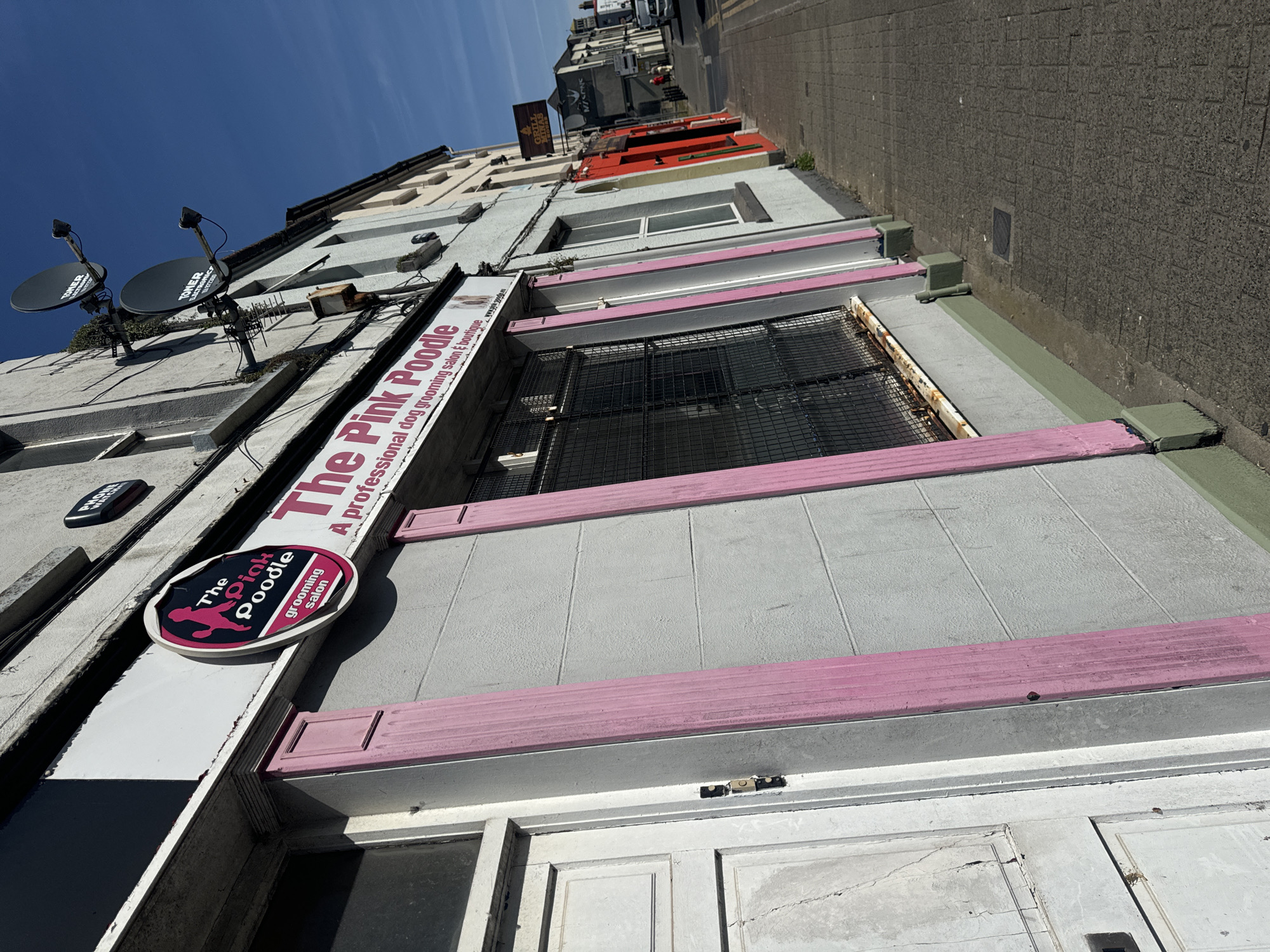 The image shows the exterior of a building with a business called "The Pink Poodle" which appears to be a professional dog grooming salon and boutique. The storefront features a prominent pink and black oval sign with a silhouette of a poodle. The building has a light grey facade with decorative pink pillars flanking the windows. A white door is visible on the left side of the image. The scene is set on a paved sidewalk in a city, with other buildings and a street visible in the background. The sky is clear and blue. Based on the presence of "The Pink Poodle" and "Grill House" signs, and the architectural style, this location is likely in Waterford City, Ireland.