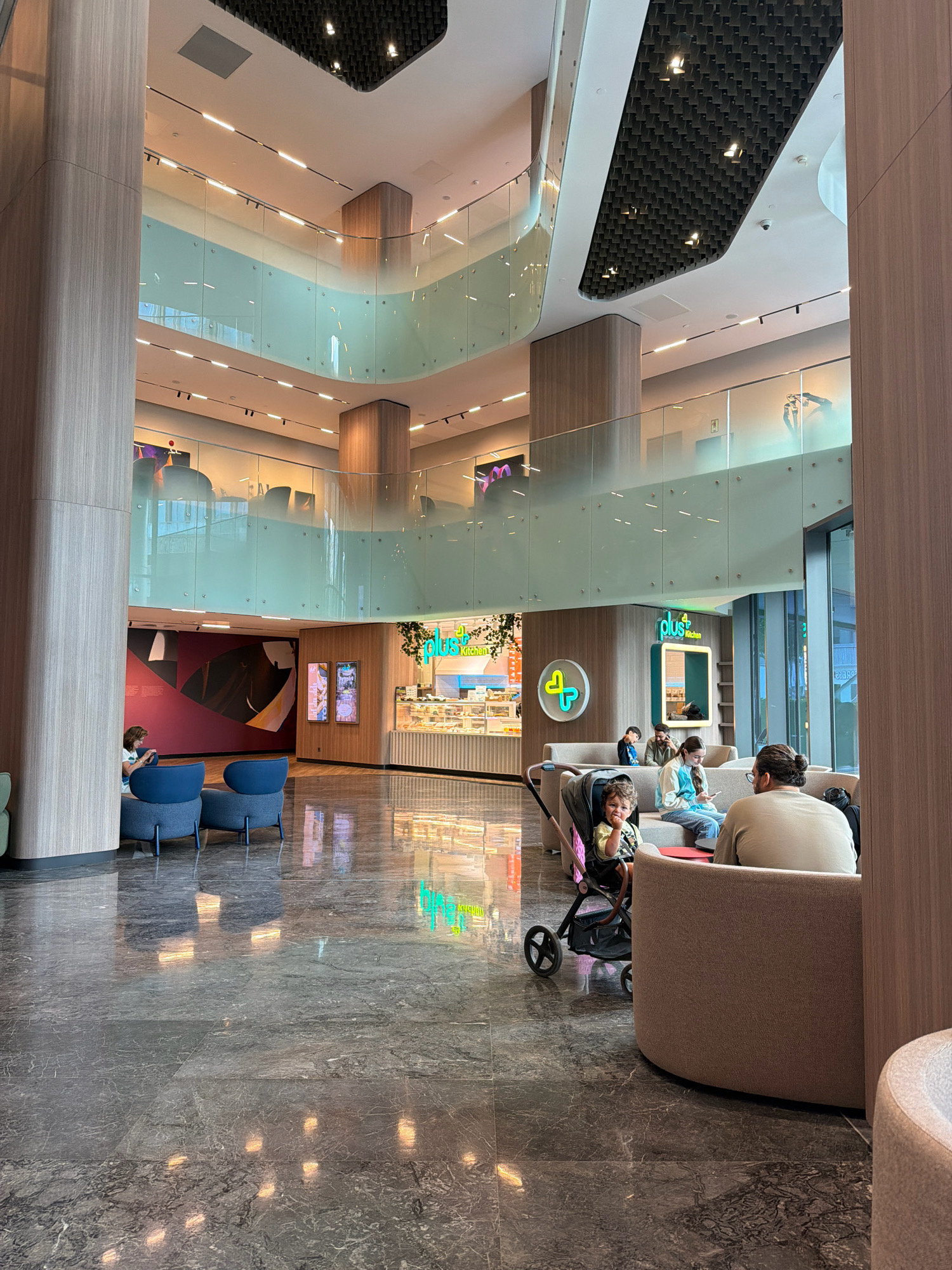 This image depicts the interior of the Palladium Tower in Ataşehir, Turkey, specifically the ground floor atrium area which features multiple levels, reflective marble flooring, and contemporary architectural design. A cafe named "plus Kitchen" is visible in the background, with displays of food and drinks.
In the foreground, several people are seated in a lounge area with rounded, upholstered seating. A woman with dark hair pulled back, wearing glasses, sits facing away from the camera on the right. In front of her, a young child in a stroller is looking towards the camera, appearing to be reaching out with their right hand. To the left of the stroller, a group of people, including a man and two women, are seated on a long sofa. One woman in the group is looking at her phone.
In the mid-ground, on the left side of the image, two blue armchairs are placed near a large wooden pillar. A woman is seated in one of these armchairs, looking down at her phone.
The atrium is characterized by its multi-story structure with glass balconies on the upper levels. The ceiling features a unique, textured pattern with integrated lighting. The overall ambiance is modern and well-lit, with natural light entering from unseen sources and artificial lighting illuminating the space. The polished marble floor reflects the light and the surroundings, contributing to the sophisticated aesthetic. The wall behind the cafe features a large abstract artwork.