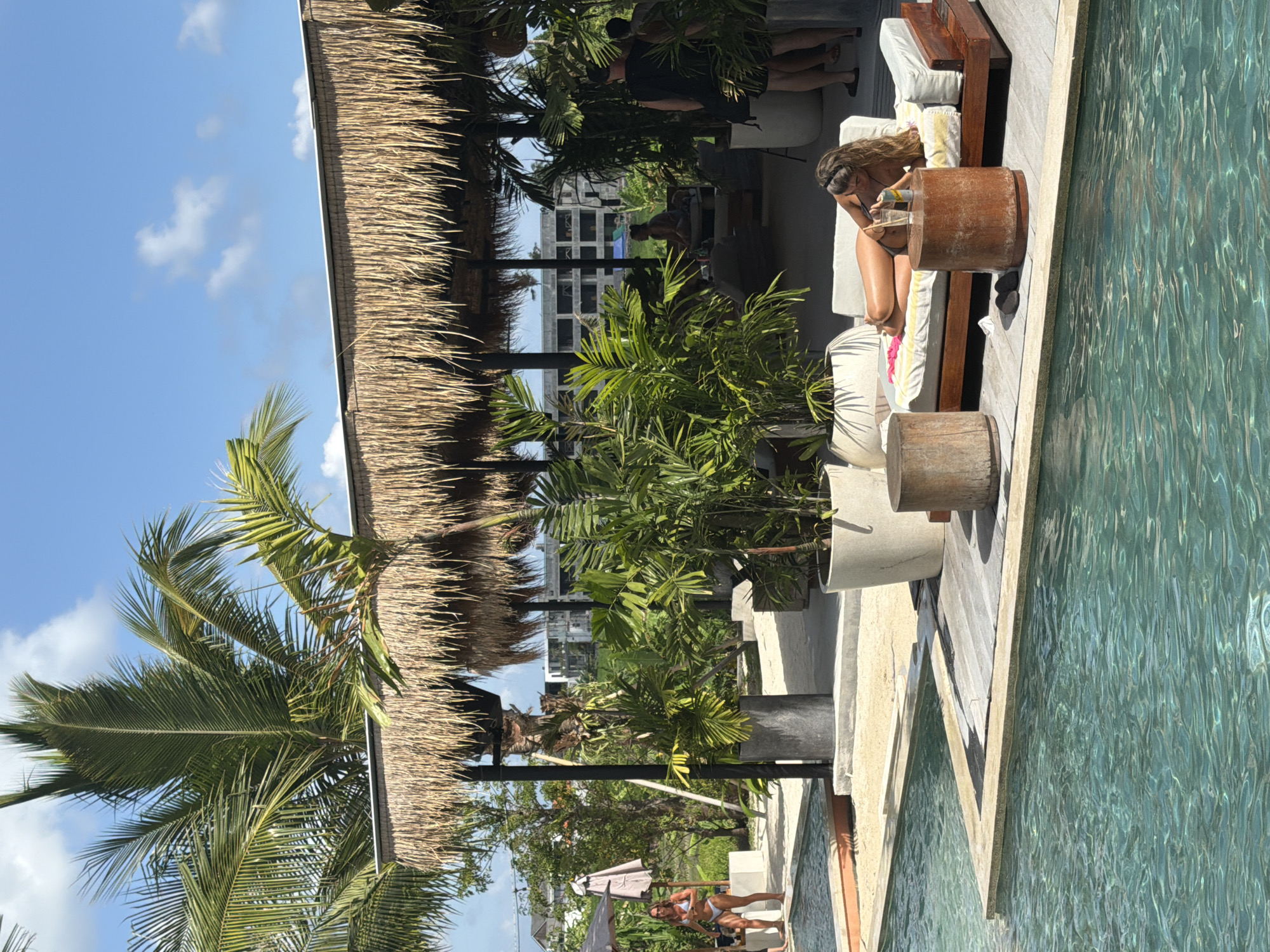 An outdoor daytime shot shows a poolside lounge area at a resort in Mengwi, Indonesia. A woman in a bikini lies on a chaise lounge by a swimming pool. She is seen from the waist up, with her head resting on a pillow and her arm bent under her head. She holds a drink in her right hand. The lounge is situated on a wooden deck with two cylindrical wooden tables in front of it. The pool in the foreground has clear turquoise water. In the background, a thatched-roof structure provides shade for an open-air seating area. Lush green plants and palm trees are visible throughout the scene, adding to the tropical ambiance. A man and woman are seen standing further back under the thatched roof. The sky is blue with scattered white clouds.