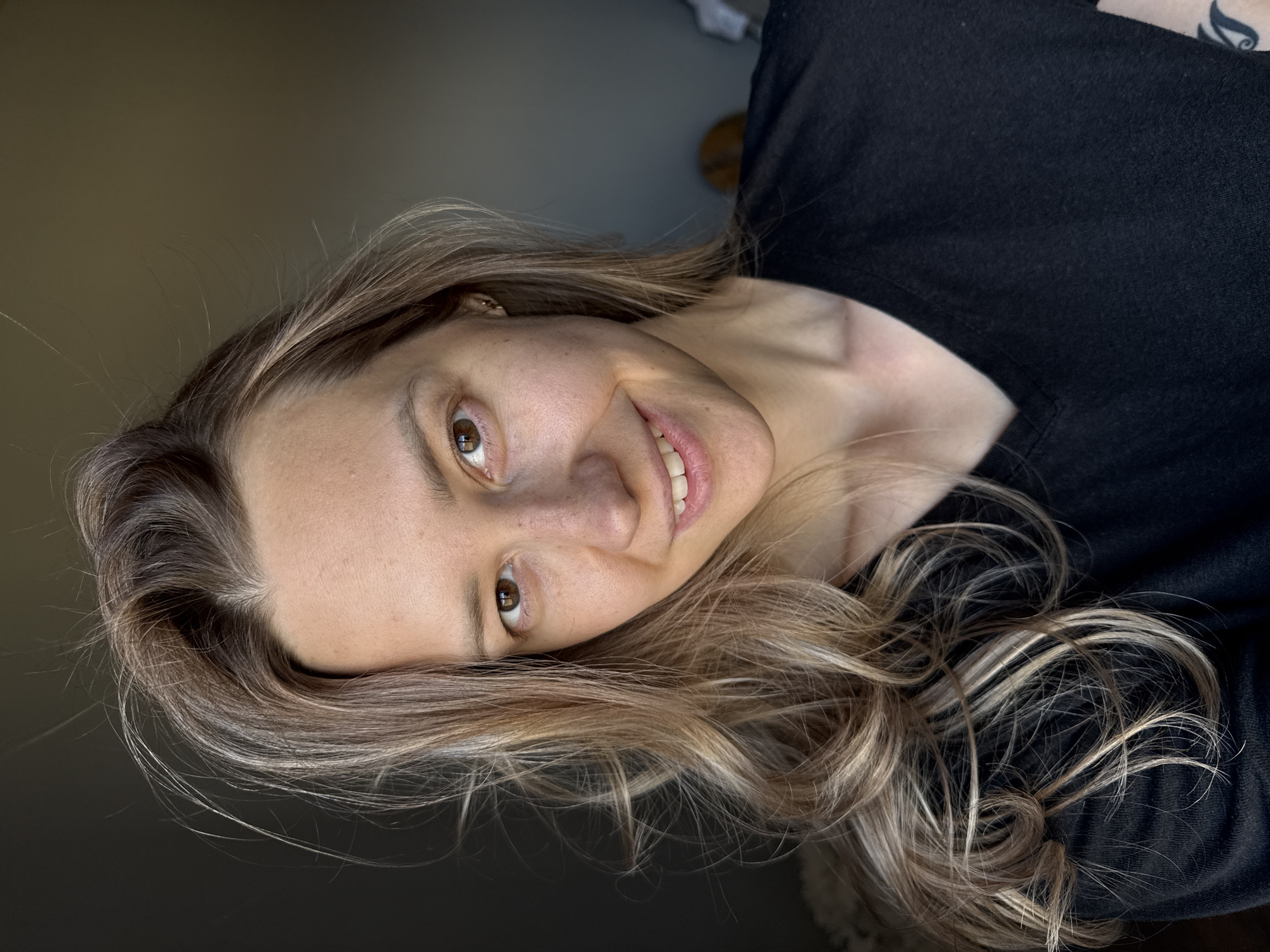 The image features a young adult woman with a natural expression, smiling gently, and looking towards the camera. Her head is tilted, giving the photo a dynamic and casual feel. She has long, wavy hair that appears to be a mix of blonde and light brown or auburn tones, styled with highlights, falling across her shoulder and chest. Her eyes are brown and appear to be looking directly at the viewer. She is wearing a simple black t-shirt or top.
The setting appears to be indoors, with a plain, blurred, dark background, likely a wall or an interior space, suggesting a soft, indirect light source illuminating her face. There are no distinct objects or features in the background that provide specific location cues, although the user context indicates the photo is from Chaikovsky, Russia.
Notable details include her clear complexion, the warmth of her smile, and the natural flow of her hair. A dark, possibly tattooed, pattern can be subtly seen on the upper part of her left arm, near the shoulder, on the right side of the frame. There is no visible text in the image. The lighting suggests it might be daytime, but there are no specific indicators of the exact time or weather. Her overall demeanor suggests contentment or happiness.