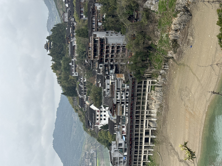 The image presents a panoramic view of a multi-tiered settlement on a steep hillside in Furong, China, under an overcast sky. A calm body of turquoise-green water occupies the lower left portion, bordered by an extensive, exposed shoreline of dry, light brown earth. This riverbed exhibits visible cracks and sedimentary patterns, indicative of low water levels. Rocky outcrops are present along the water's edge and at the base of the hillside structures.

The town consists of numerous multi-story buildings clustered densely on the hillside. Architectural styles predominantly feature dark tiled roofs and light-colored facades, with some displaying traditional Chinese elements like ornate balconies and layered eaves. The lowest tier of buildings, immediately above the exposed shoreline, comprises a long, multi-level structure supported by multiple concrete columns and beams, forming an elevated promenade or foundation. Red lanterns are suspended from railings on these lower levels. Higher on the slope, buildings are integrated with the natural terrain and interspersed with dense green vegetation. At the highest visible point of the central hill within the town, a prominent, multi-tiered traditional Chinese pagoda-style building with a dark roof is situated amidst trees.

One individual, dressed in blue, is observed on the exposed dry riverbed near the lower right rocky bank. In the far background to the left, a large grey infrastructure complex, possibly a bridge or dam section, borders the water, set against a backdrop of distant green mountains. The overall environment suggests a mountainous riverine setting.