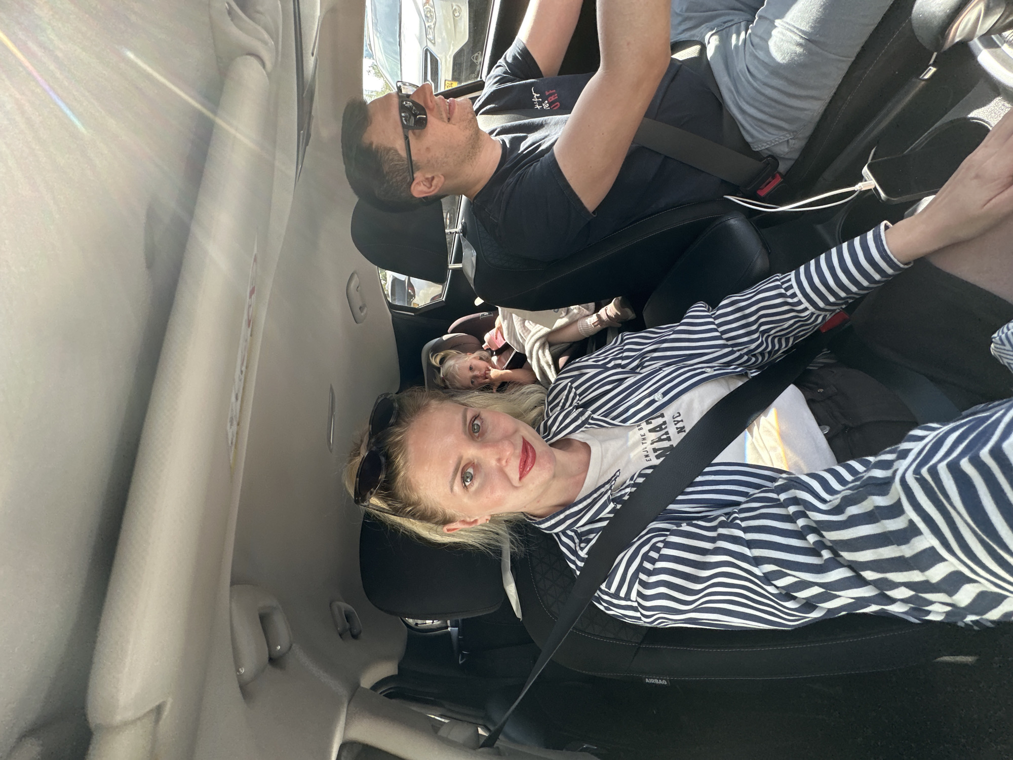 Inside a vehicle, a fair-skinned woman with blonde hair and red lipstick, wearing a white t-shirt with "ENJOY THE BR.... NYC" text and a blue and white striped shirt, is in the passenger seat. She has sunglasses on her head. A man with sunglasses, wearing a dark t-shirt, is in the driver's seat. A small child is lying in the back. The vehicle is likely in motion, based on the blurry outside view. The interior of the vehicle is visible with a focus on the occupants. The location is Petah Tikva, Israel.