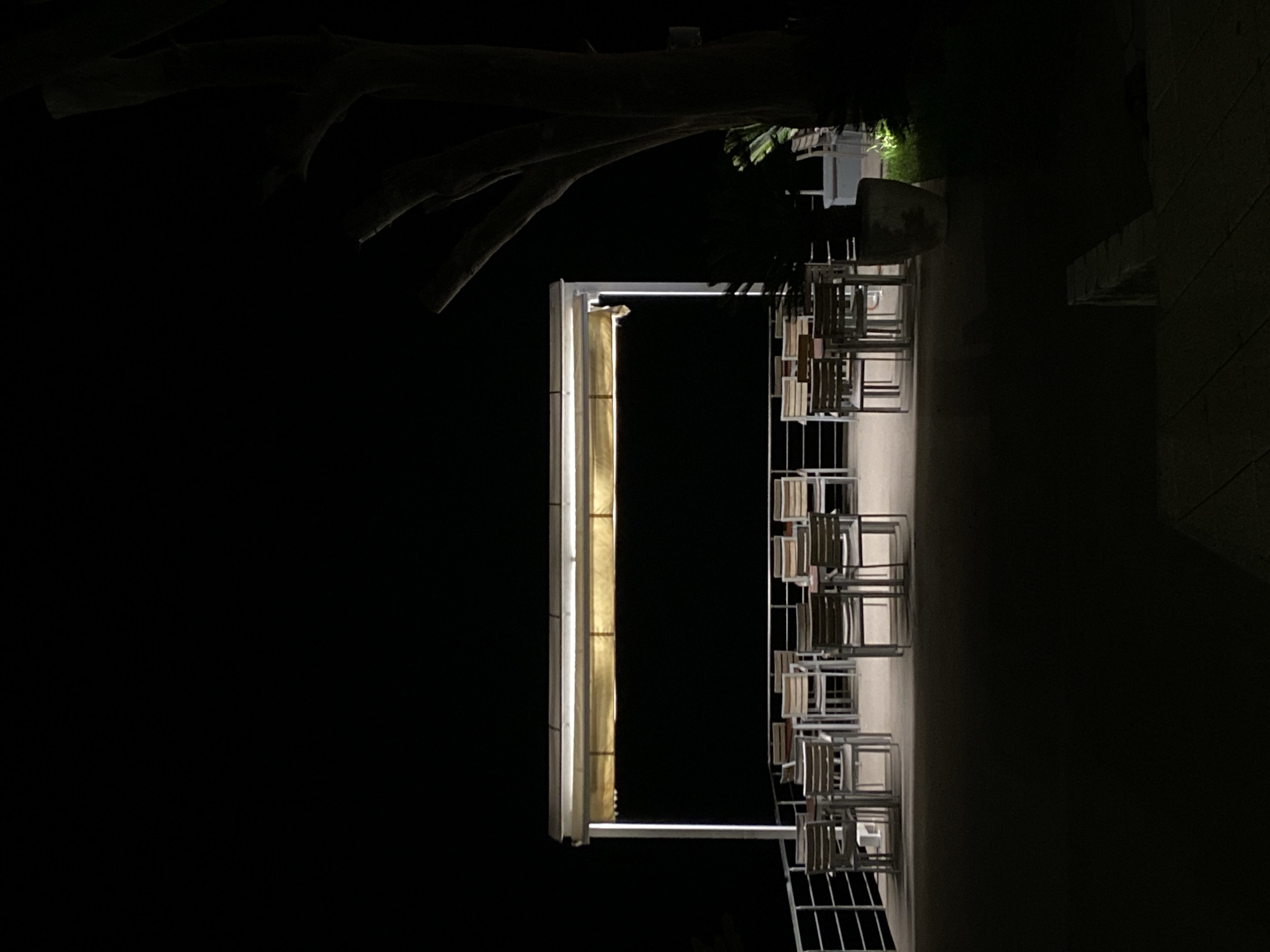 The image depicts an outdoor scene at night, possibly in or near Bar, Montenegro, showing a structure with a bright, illuminated vertical element. Adjacent to this, there are stacks of chairs, seemingly for an outdoor dining or seating area. A large tree branch is visible in the top left corner, contrasting against the dark sky. To the right, there's a wall-like structure or the side of a building. The overall lighting is dim except for the bright vertical light, creating stark shadows and emphasizing the shapes and forms in the scene.