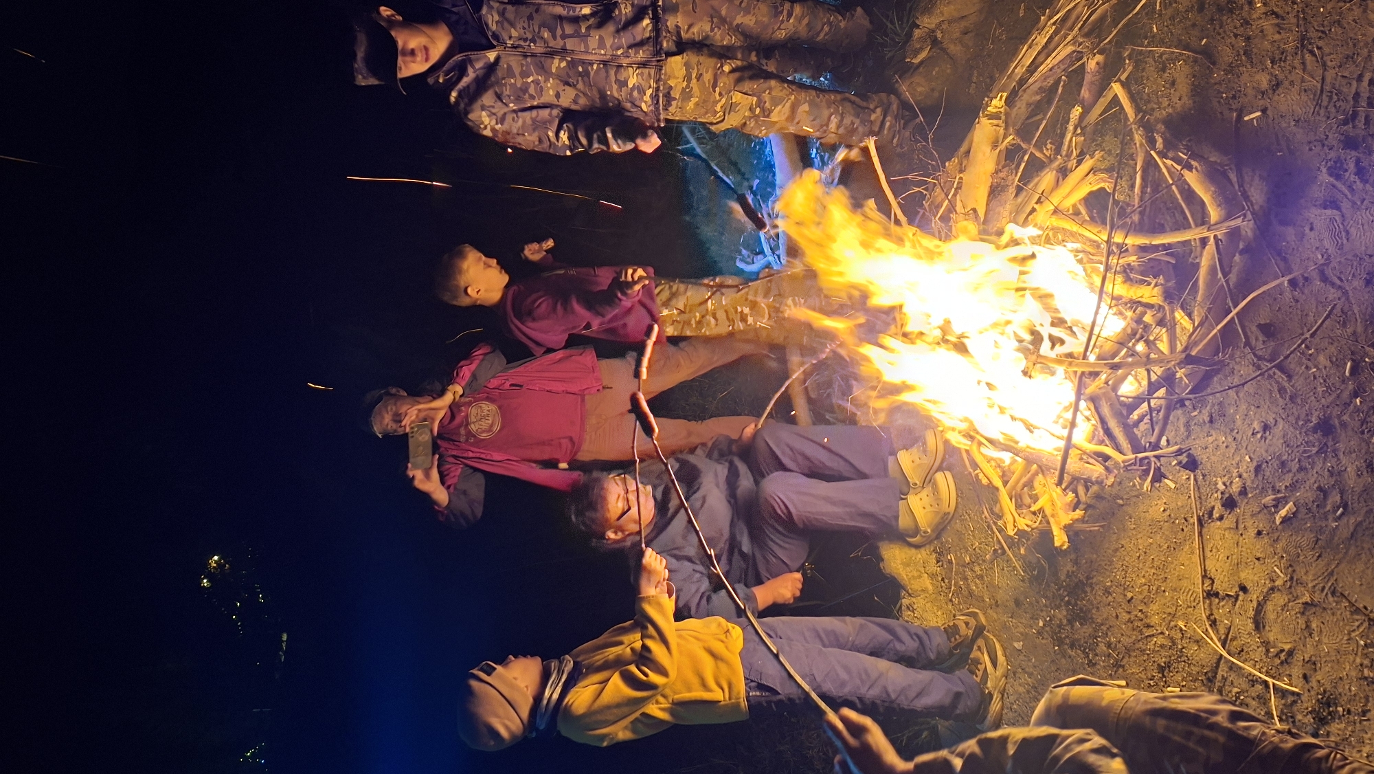 The image depicts a nighttime outdoor gathering around a bonfire, likely in Batan, Kazakhstan, though the specific location is not definitively identifiable. Five individuals are visible. In the foreground and to the left, a young person wearing a yellow jacket and a light-colored beanie holds a stick with food over the fire. To their right, an adult in a dark blue jumpsuit and light-colored clogs is seated, also with a stick and food over the fire. Further back, to the left, an individual in a pink jacket is holding up a mobile phone, appearing to take a photo or video. Next to them, a young person in a purple hoodie and camouflage pants stands observing the fire. On the far right, another individual in a camouflage jacket and cap is present. The bonfire is the central focal point, with bright flames illuminating the faces and clothing of those around it. The ground appears to be dirt or sand, and the background is dark, suggesting nighttime. Some sparks are visible rising from the fire, and the overall atmosphere suggests a casual, communal activity.