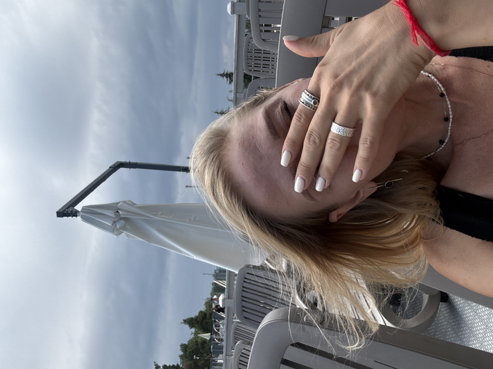 A woman is partially visible in a close-up shot, covering her right eye with her hand. She has blonde hair, white-painted fingernails, and is wearing several rings on her left hand. She also has a necklace with pearls and black beads, and a red string bracelet on her right wrist. In the background, there are rows of empty gray lounge chairs, a white umbrella, trees, and a cloudy sky. The overall scene suggests an outdoor seating area, possibly at a resort or waterfront establishment. The location is not explicitly shown or identifiable as Yalta, Ukraine.