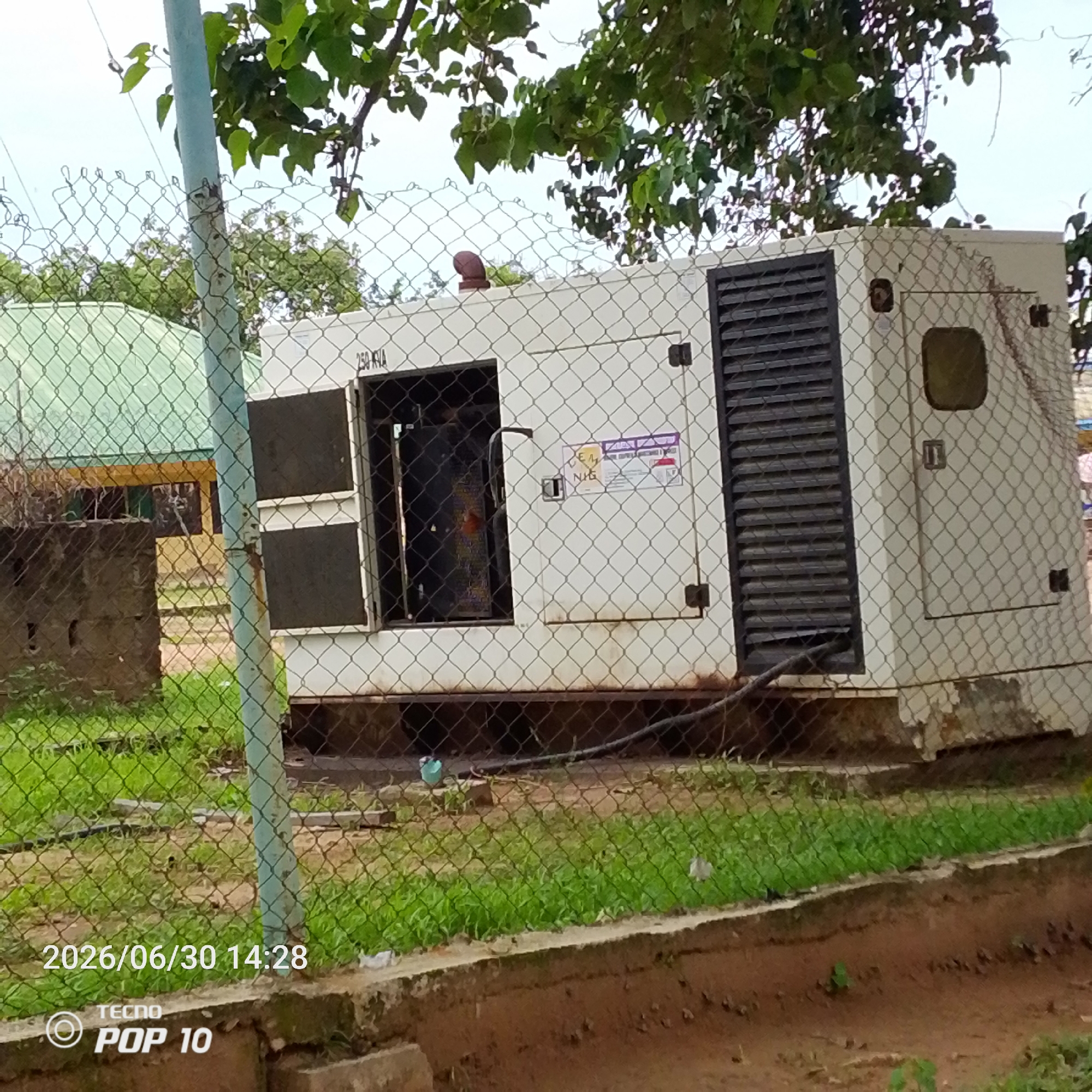 This image depicts an outdoor scene featuring a large white generator enclosed by a chain-link fence. The generator appears to be a portable unit, with a dark grille on its right side and an open panel on its left, revealing internal components. A sticker with text and a yellow circular emblem is affixed to the generator's door. The generator is situated on a concrete base, and the surrounding ground is covered in green grass and some patches of dirt.
In the background, a building with a green roof and yellow walls is partially visible, suggesting an institutional or commercial setting. A concrete structure, possibly a wall or planter, is also present to the left of the generator. Overhanging tree branches with green leaves are seen at the top of the image, indicating an outdoor environment.
The scene is captured on a bright day, likely daytime, given the lighting. The presence of the generator suggests a location that may experience power outages or requires a backup power source. The timestamp "2026/06/30 14:28" is overlaid on the image, along with "TECNO POP 10," indicating the date and time of capture and the device used. There is no visible indication of people or specific activities occurring at this moment, leading to a static, observational portrayal of the generator and its surroundings in Jalingo, Nigeria.