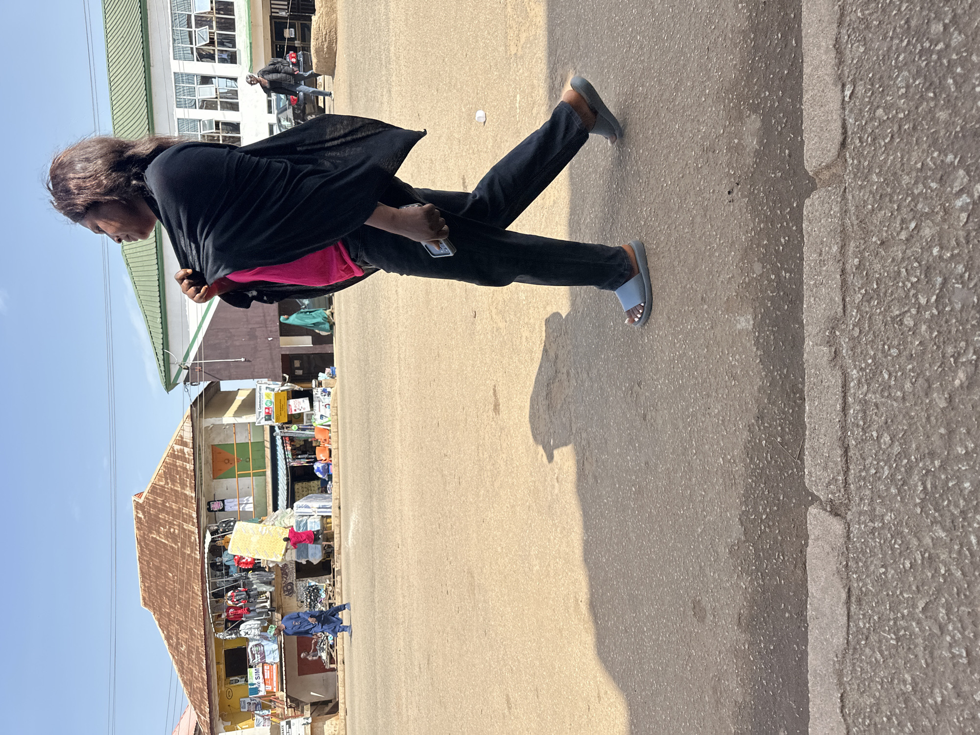 A woman walks across a dusty, paved street in Bukuru, Nigeria. She is wearing a black shawl, a pink shirt, and black pants, with light blue slide sandals on her feet. She holds a mobile phone in her right hand. In the background, a two-story building with a corrugated metal roof is visible, housing several small shops displaying clothing and other merchandise. To the right, another building with a green roof and large windows is also present. Several other people are walking in the distance, and a car is parked near the green-roofed building. The sky is clear and blue.