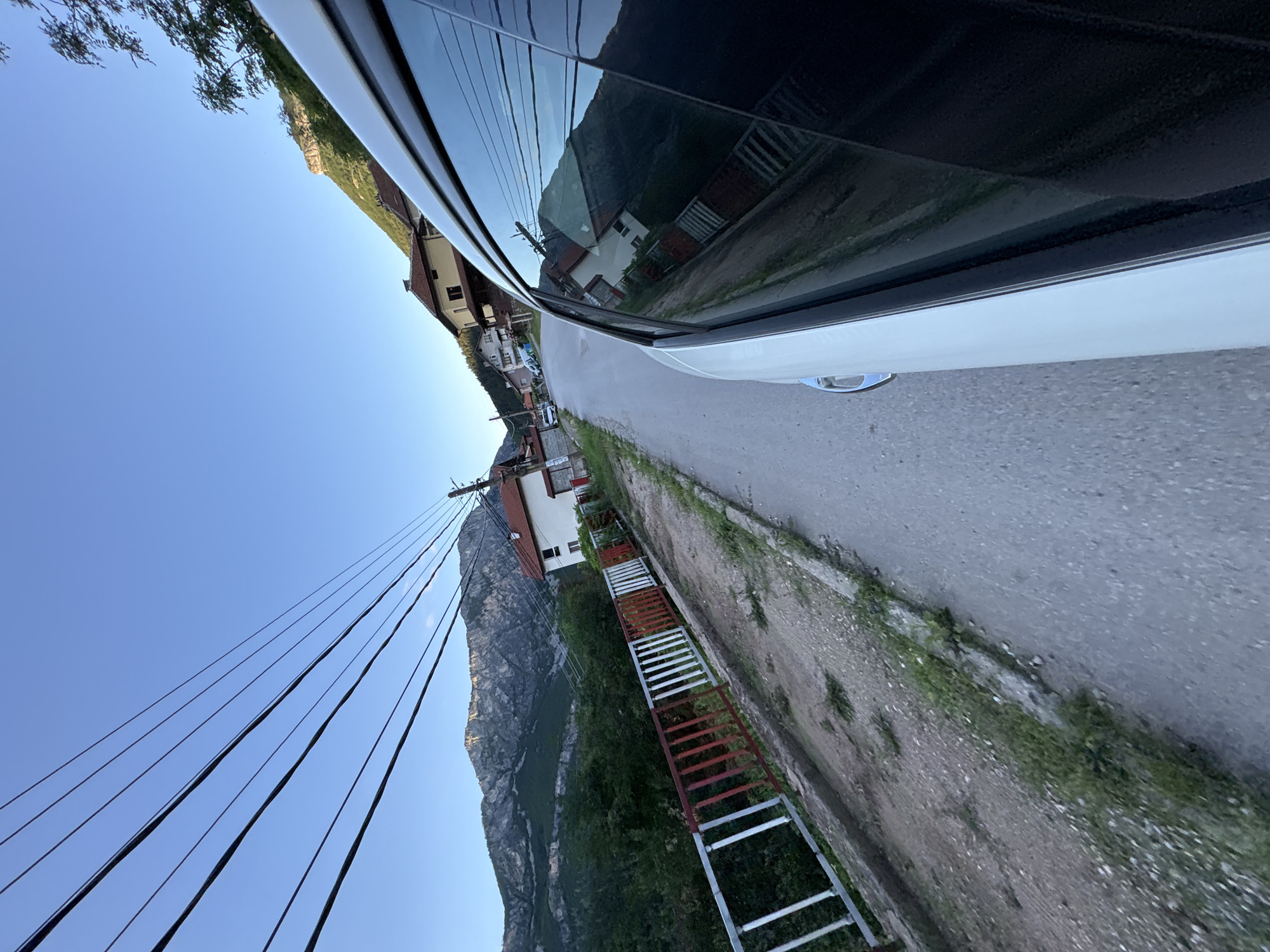 The image captures a street scene in Zgorigrad, Bulgaria. A white vehicle is positioned on the right side of the road, its side and tinted window dominating the foreground. The road is paved and slopes gently uphill. To the left of the road, a metal fence with red and white bars runs along a dirt embankment. Beyond the fence, dense green trees line the landscape. In the background, houses with red-tiled roofs are visible nestled amongst the trees and along the hillside. Towering over the town are rugged, tree-covered mountains under a clear blue sky. Overhead, multiple power lines stretch across the frame, suspended from utility poles. The lighting suggests it is either early morning or late afternoon.