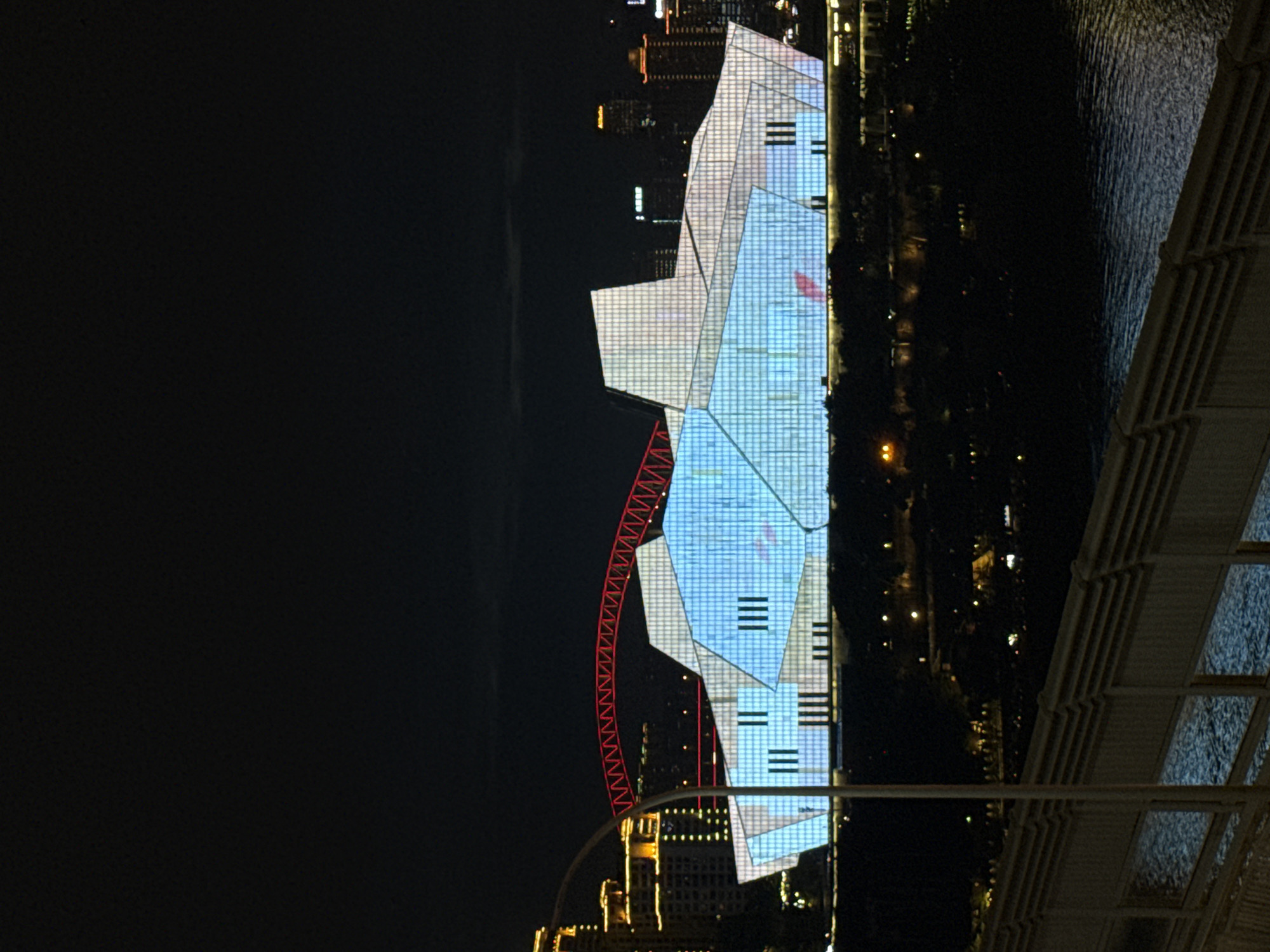 The image shows a modern building with a glass facade illuminated with blue and white lights at night. The building has a geometric, angular design. A red, arched bridge is visible behind the building. In the background, city lights and taller buildings can be seen, indicating an urban environment. In the foreground, a body of water is visible, with a railing or wall along its edge. The overall scene suggests a city skyline at night, likely in a developed area. The building's illumination suggests it is a prominent landmark. The location is identified as Yuzhong District, China.