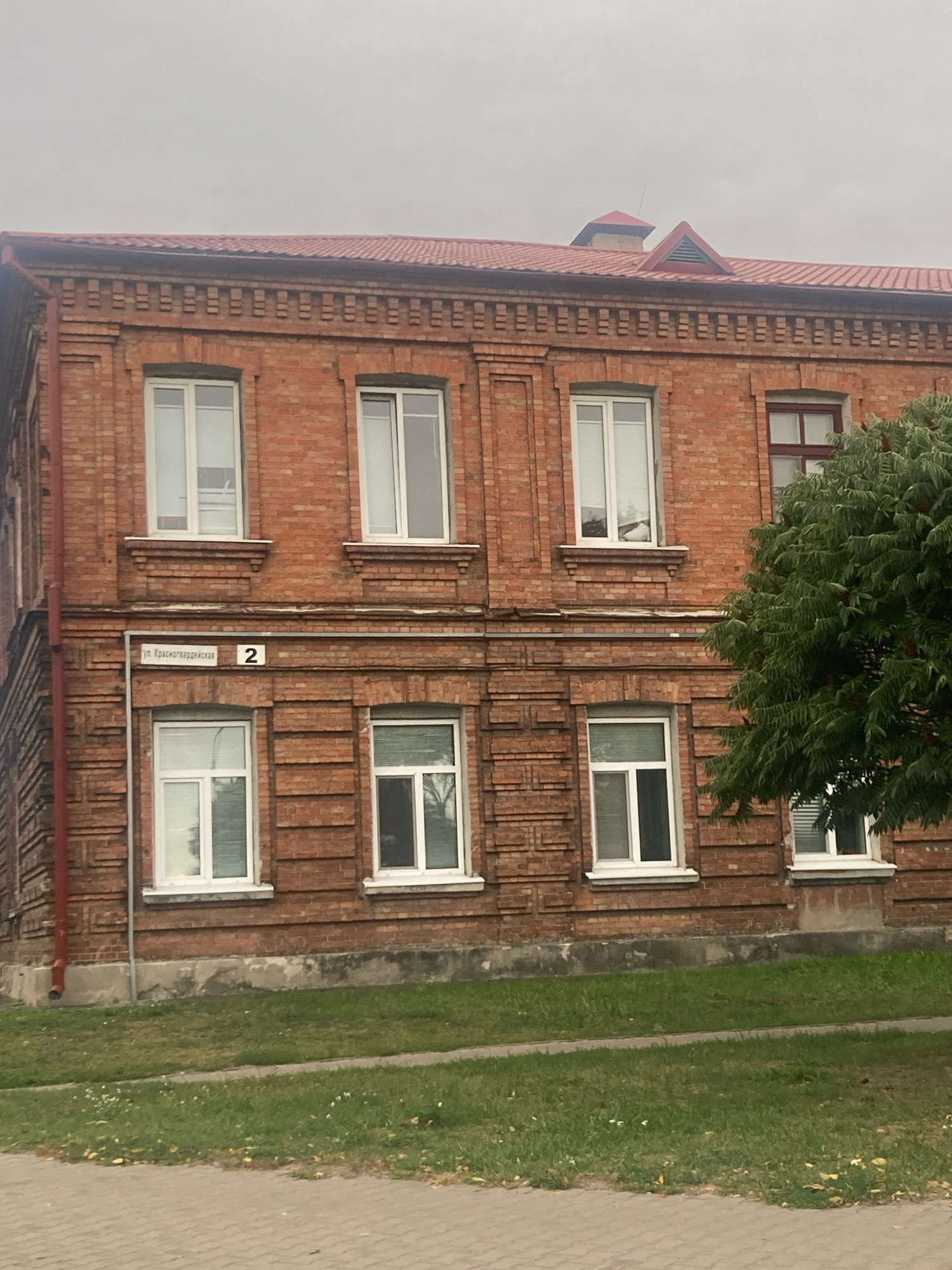 This image depicts a two-story brick building with a red tiled roof, located in Brest, Belarus. A street sign on the building reads "ул. Красногвардейская 2" which translates to "Krasnogvardeyskaya Street 2". The building has multiple windows with white frames. The lower section of the building appears to be a basement or foundation. In front of the building, there is a grassy area with a paved walkway. A large green tree is visible on the right side of the image, partially obscuring some of the building's windows. The sky is overcast. There are no people visible in the image. The scene is a static exterior shot of an urban environment.
