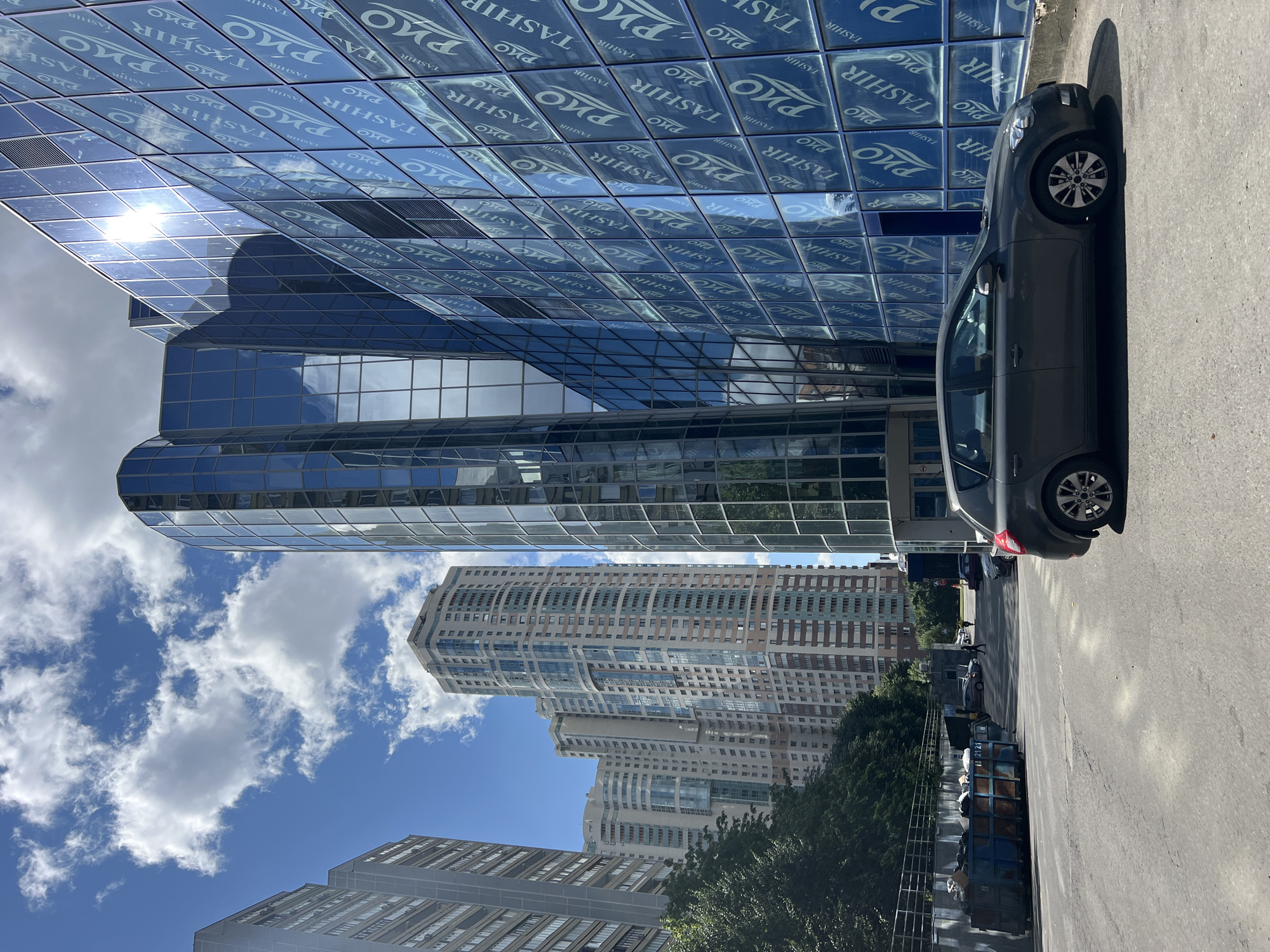 This is a street-level, eye-level, eye-level, medium shot of a modern urban environment in Moscow, Russia. The image features a prominent, modern building with a glass facade displaying the "TASHIR" logo repeatedly, reflecting the blue sky and white clouds. To the left of this building, several tall residential high-rises are visible. In the foreground, a dark gray hatchback car is parked on an asphalt surface. Further down the street, other vehicles and pedestrians are present, along with large industrial trash containers. The overall scene depicts a sunny day in a bustling city district.