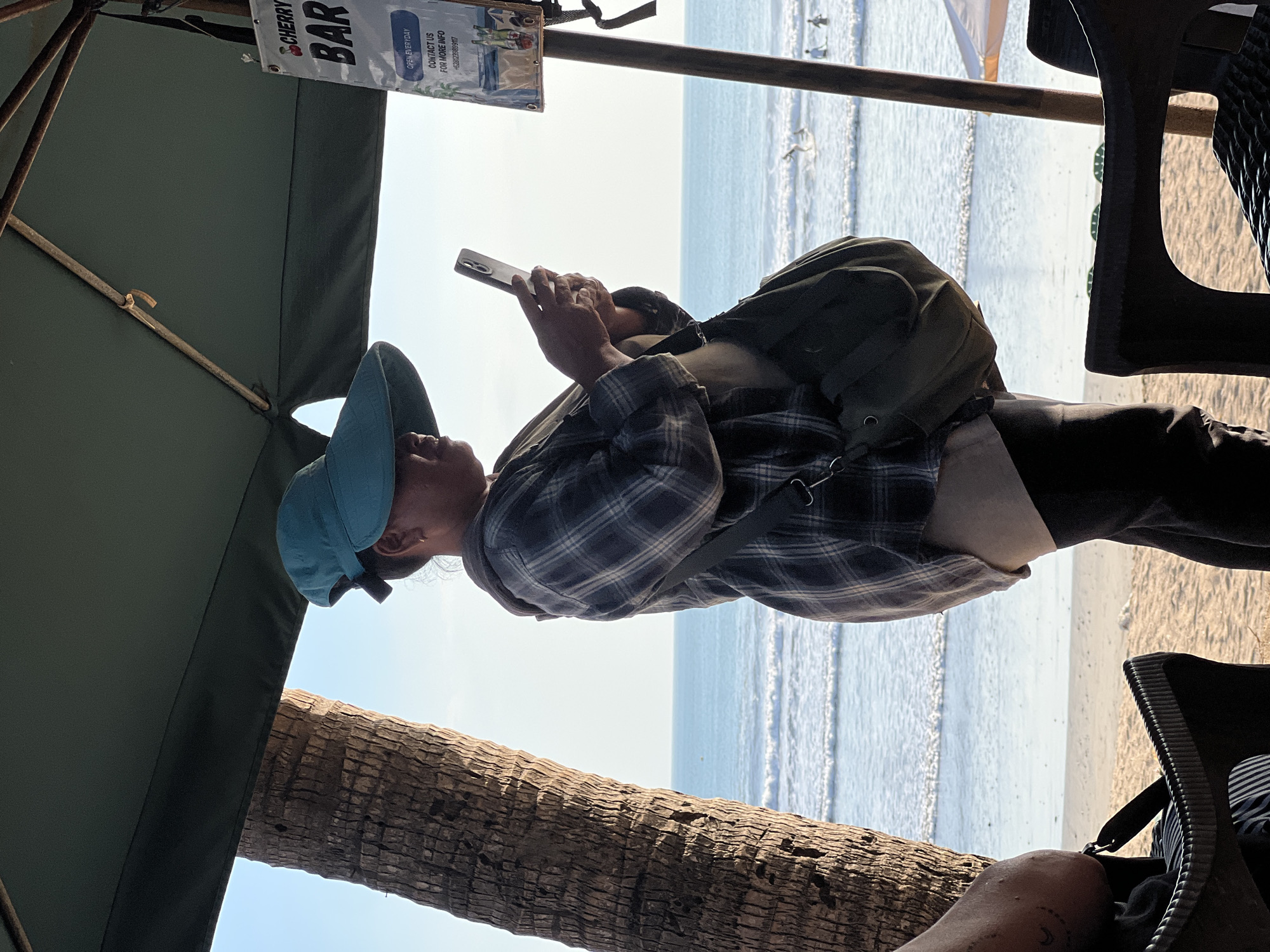 A woman wearing a large, wide-brimmed turquoise hat stands on a beach in Kuta, Indonesia, holding a smartphone in her hands. She is looking down at the phone, which is held in landscape orientation. She is wearing a plaid long-sleeved shirt over a lighter colored garment and dark pants. A dark green bag is slung across her body. A palm tree trunk is visible to her left, and an umbrella provides shade above her. In the background, the ocean stretches out with gentle waves, and several people can be seen swimming or surfing in the water. A sign for "CHERRY BAR" is attached to a wooden post on the right side of the image. The scene appears to be during the daytime with clear skies.
