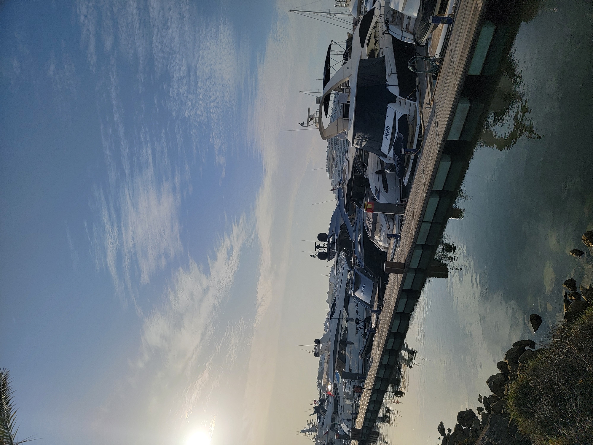 The image shows a marina in Quarteira, Portugal, filled with several luxury yachts docked along a wooden pier. The sun is low in the sky, casting a warm glow on the scene and creating reflections in the calm water. The sky is a clear blue with scattered white clouds. The foreground features a rocky shoreline with some vegetation. The yachts are of various sizes and designs, with some covered by tarpaulins. The overall atmosphere is one of tranquility and leisure.