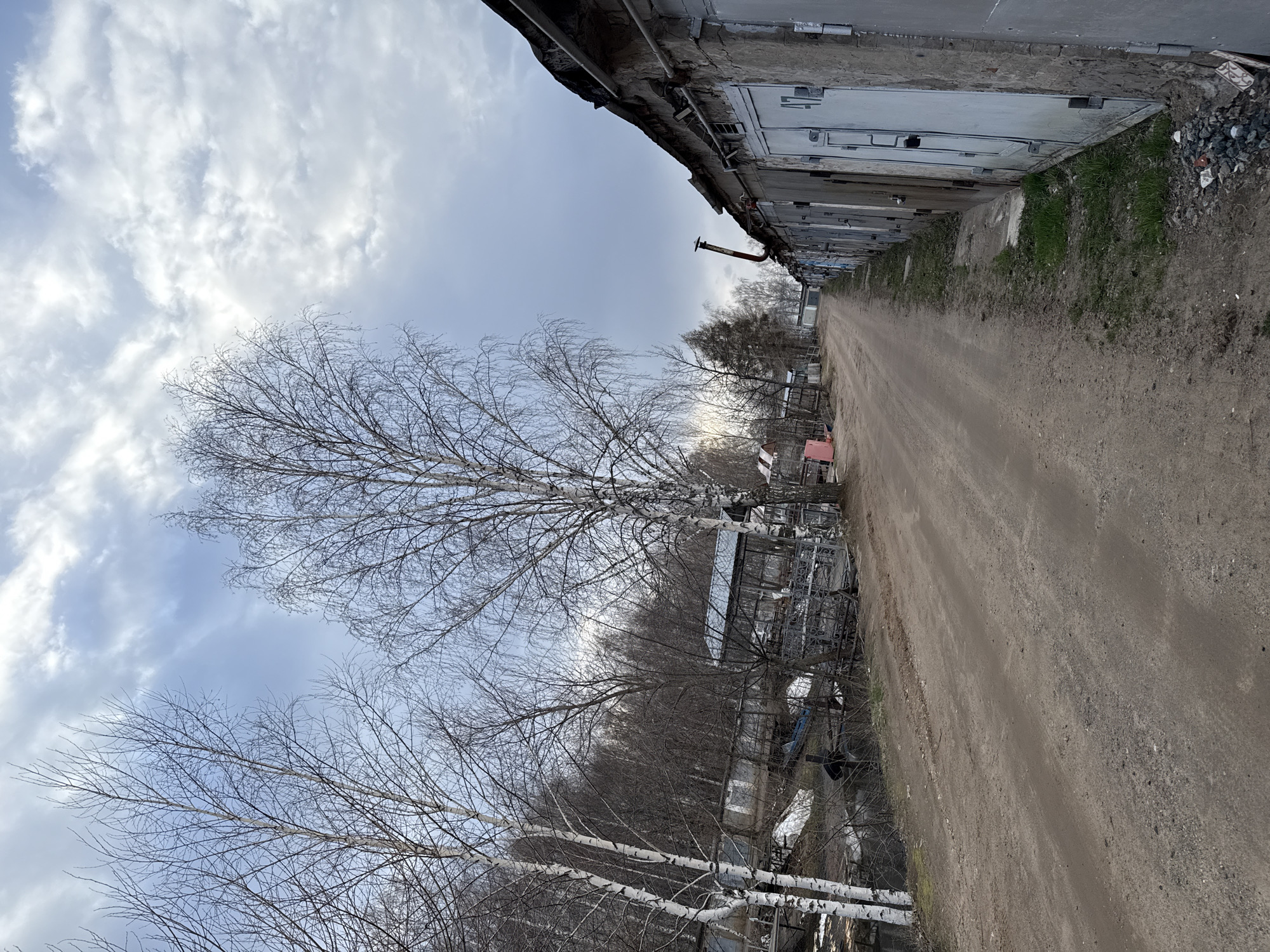 A ground-level, outdoor shot of an alleyway or narrow dirt road in an urban area of the city of Unknown, Unknown. The alley is flanked by rows of garages on the right side of the frame. These garages are constructed with grey or white siding and metal doors, their facades showing varying degrees of weathering and wear. The left side of the alley features a row of bare trees, birch trees notable among them, with their branches stark against a sky filled with white and grey clouds. The dirt road itself is uneven and appears lightly traveled, with some sparse grass and vegetation growing near the edges of the alleyway. In the middle distance, there is what looks like metal frames, or other urban elements along the side of the alley. There are no people visible in the scene. The overcast sky suggests a muted light across the environment.