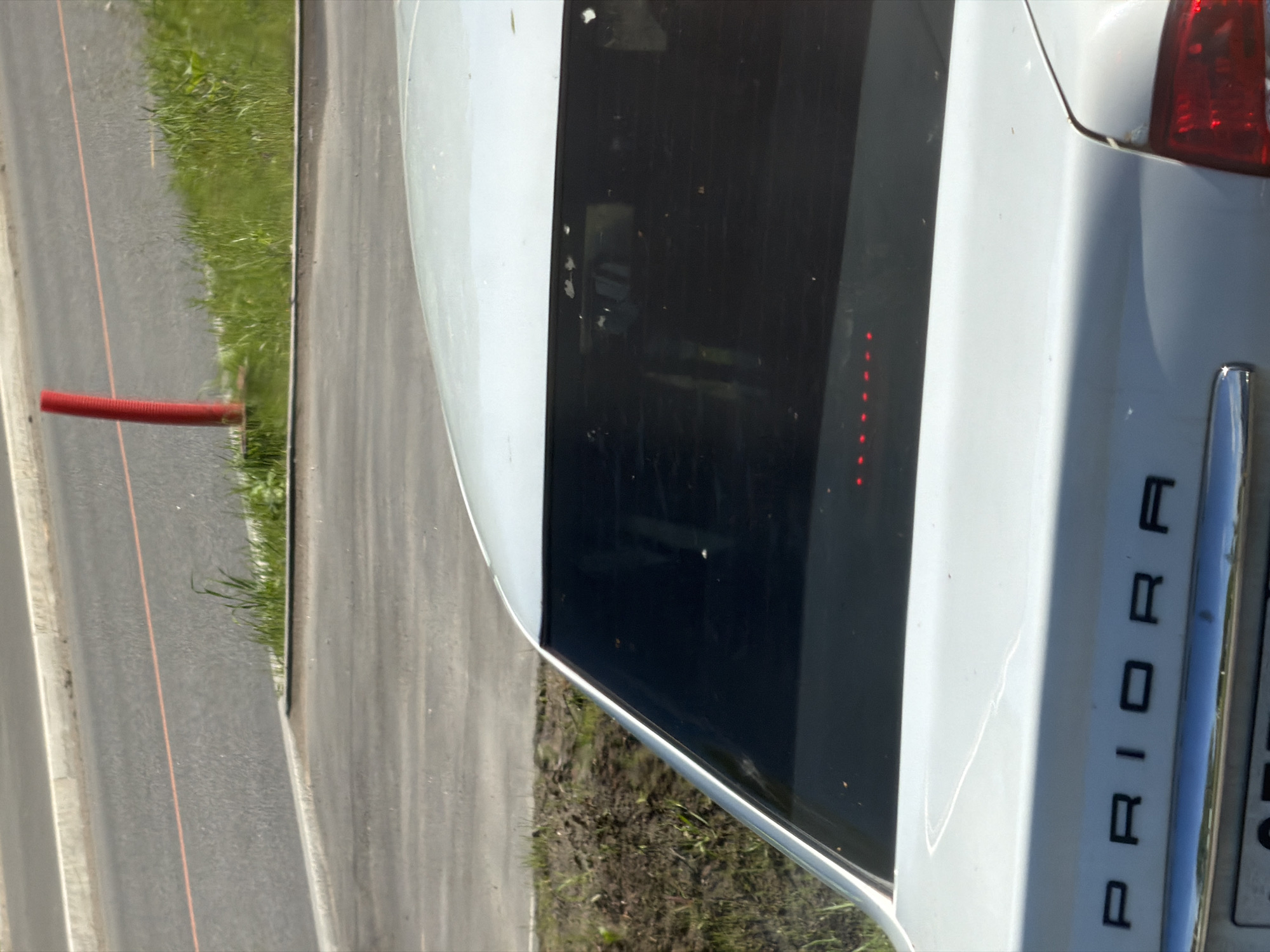 The image shows the rear of a white Lada Priora vehicle, with "PRIORA" spelled out in capital letters vertically along the right-hand side of the trunk. The rear window is heavily tinted. The vehicle is parked on a paved surface, with grass and a roadside ditch visible to the left. The red object that is visible to the left of the photo suggests a road and possibly roadworks. The location is likely in Yanino-1, Russia. No people are visible in the image.