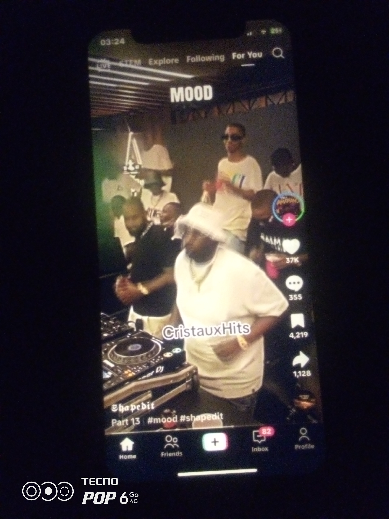 The image is a screenshot of a TikTok video playing on a smartphone. The video displays a group of people at what appears to be a music event. In the foreground, a person in a white t-shirt and headscarf is visible, standing in front of a DJ setup. Another person in a black t-shirt is also near the DJ equipment. Several other individuals are in the background, some dancing and others observing. The video interface elements include a time stamp "03:24", navigation options like "LIVE", "STEM", "Explore", "Following", and "For You", as well as icons for likes, comments, shares, and profile. Text overlays on the video read "MOOD", "CristauxHits", "@shajedit", and "Part 13 | #mood #shajedit". The bottom of the screenshot shows the TECNO POP 6 4G branding. There is no information in the image that indicates the location is in the city of Howayi, Nigeria.