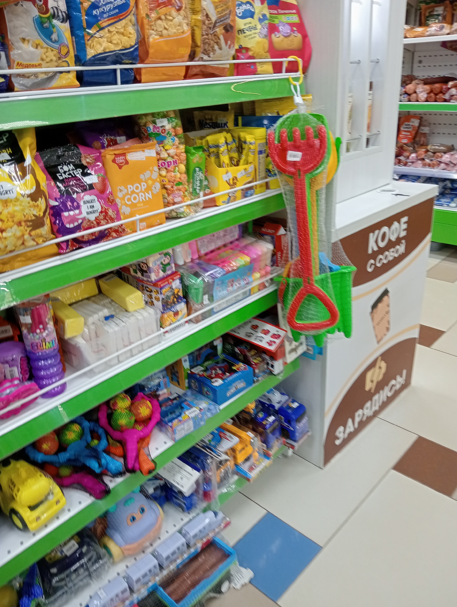 The image shows a retail store aisle with shelves stocked with various products, primarily children's toys and snacks. The shelves are green and white. On the top shelf, there are bags of cereal and snacks with Russian text. On the second shelf, there are more bags of snacks, including popcorn. A set of colorful plastic beach toys, including a rake and shovel, are hanging on a hook. The third shelf contains children's toys, including what appear to be slime containers and building blocks, as well as some packaged goods. The fourth shelf displays more toys, including toy cars and what looks like a stuffed toy animal. On the bottom shelves, there are various toys, including toy trains and what appears to be a remote-controlled car. To the right of the shelves, there is a white display unit with brown text that reads "КОФЕ С СОБОЙ" (Coffee to go) and "ЗАРЯДИСЬ!" (Charge up!). The floor is tiled with a pattern of white, beige, and brown tiles, with a blue tile visible in the lower left corner. There are no people visible in the image.