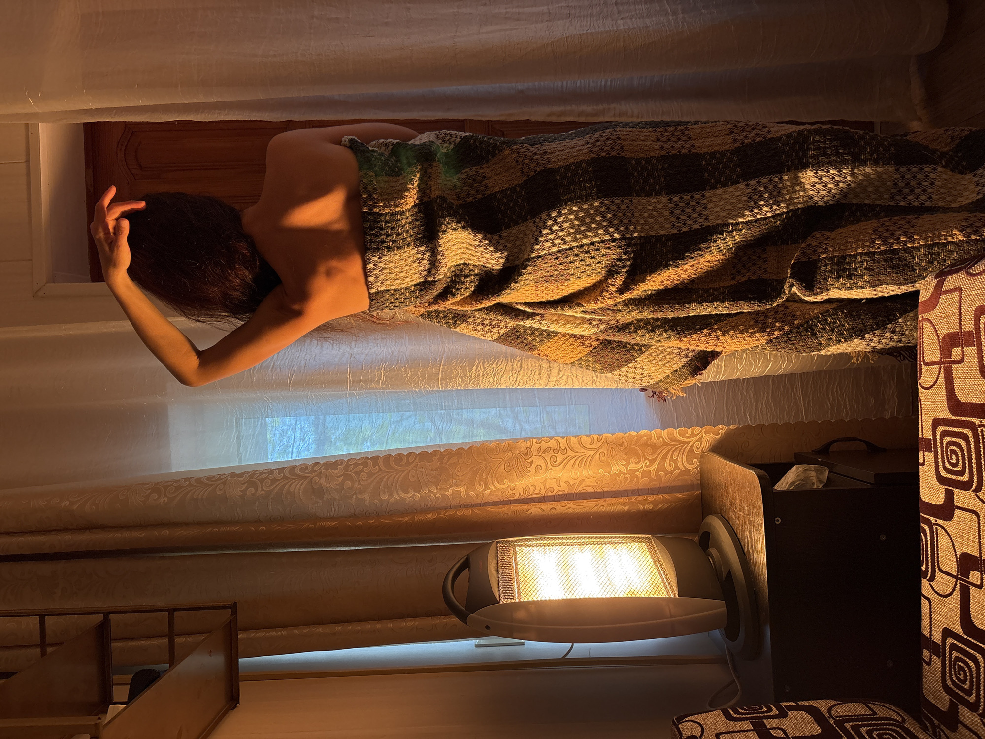 A partially nude, dark-haired person lies under a brown, patterned blanket in what appears to be a bedroom. The person's back is visible, and one arm is raised near their head. The room has cream-colored curtains, a space heater, and various pieces of furniture. The location is Gelendzhik, Russia.