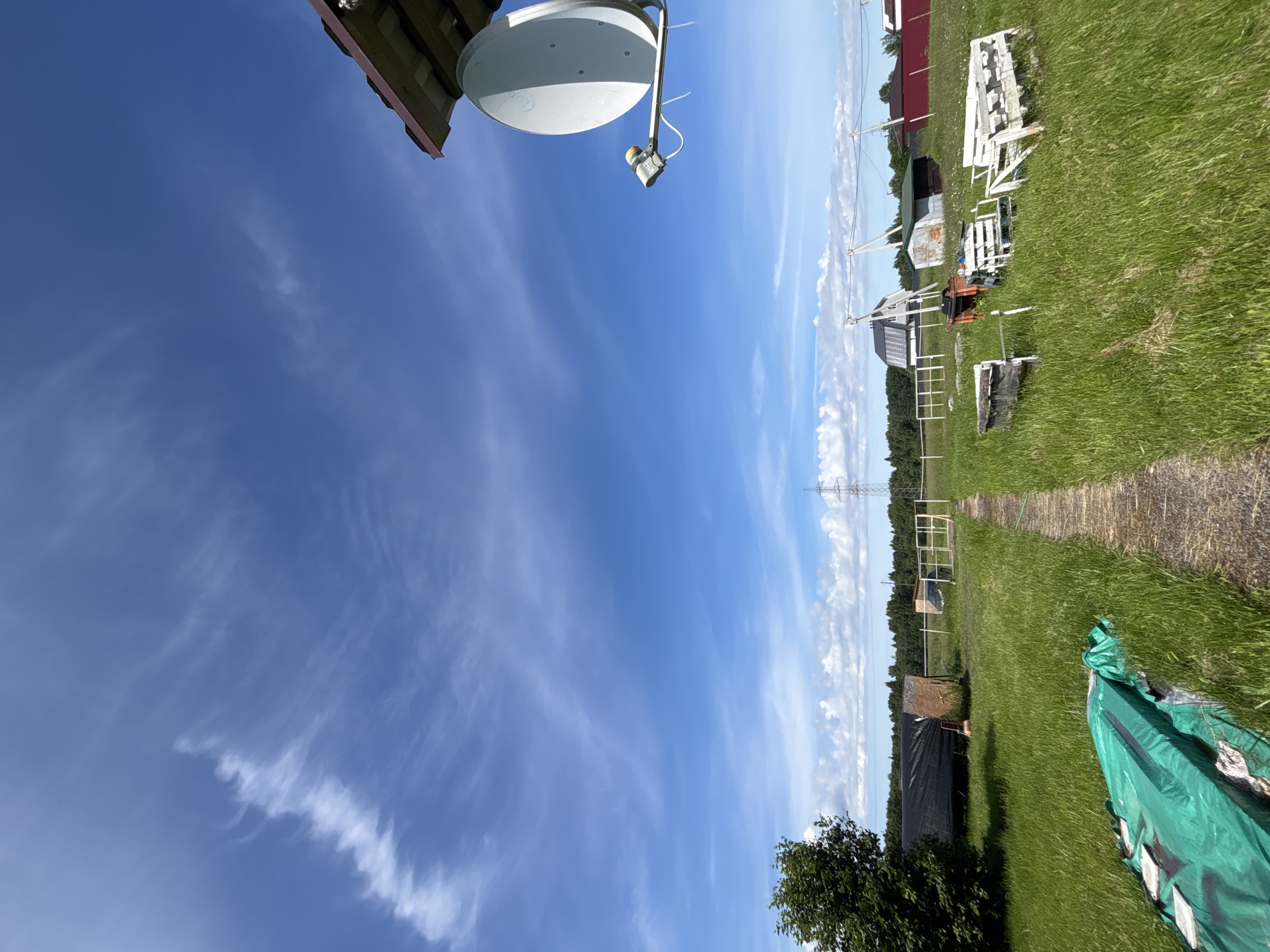 The image depicts a rural outdoor scene under a bright blue sky with wispy white clouds. The foreground features a narrow dirt path leading into a grassy field. To the left of the path, a large green tarp is spread out, partially covering some unseen objects. In the middle ground, several wooden structures, possibly benches or old farm equipment, are scattered in the grass. Behind these, a long, dark-covered structure, likely a greenhouse or storage shed, stands to the left. Further back, a fence runs horizontally across the field, with several buildings visible beyond it, including a house with a modern solar panel roof. A tall transmission tower is visible in the distance. On the right side of the image, attached to the eaves of a building, is a satellite dish. The environment is lush with green grass and trees, suggesting a rural or suburban setting in the city of Razdolye, Russia. There are no people visible in the image.