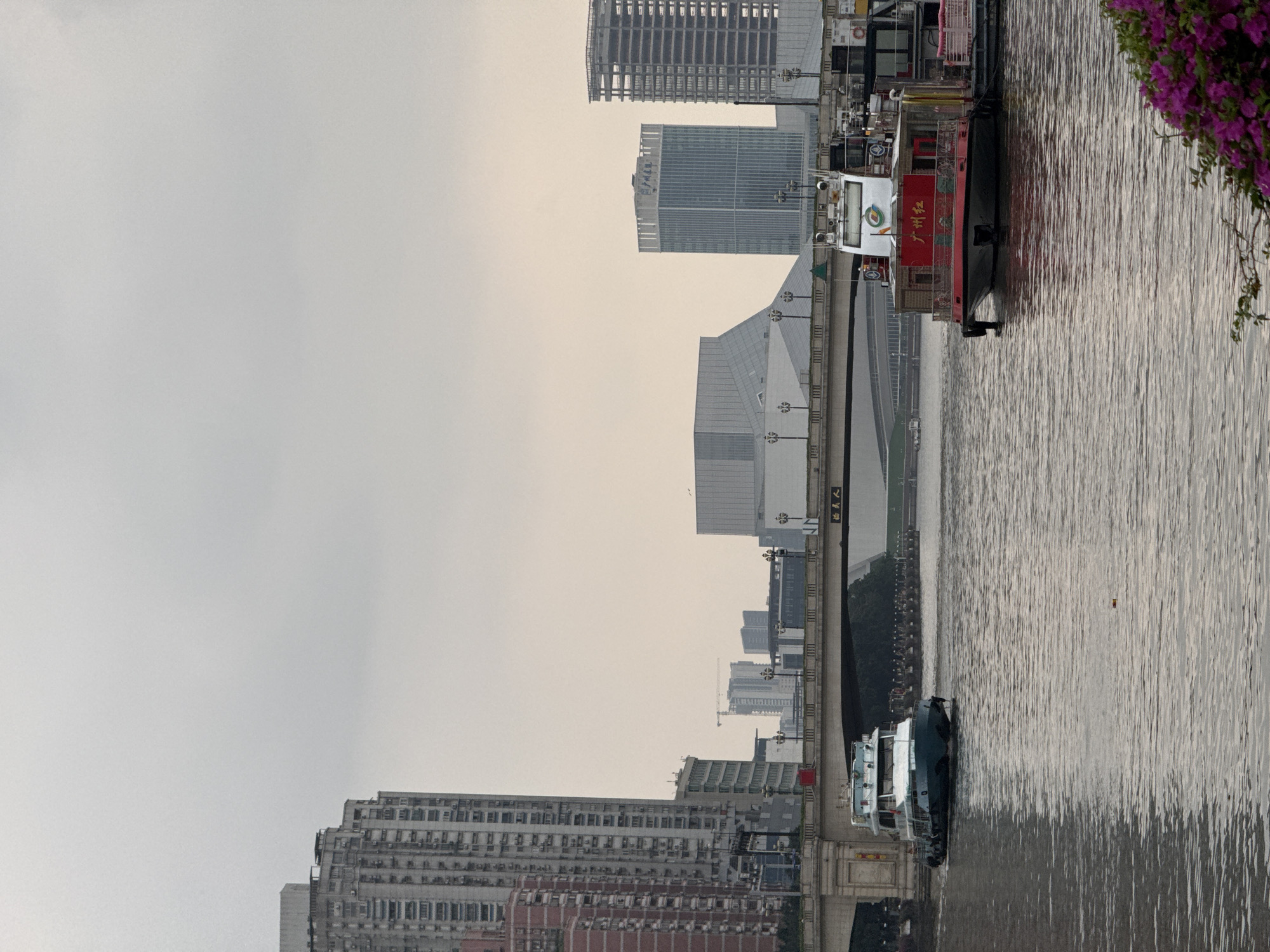 The image shows a waterfront view in Yuexiu District, China. The camera faces a waterway, with boats docked. A variety of buildings are visible on the opposite bank, including apartment buildings and office towers. A bridge is also visible, running over the river. The sky is overcast.