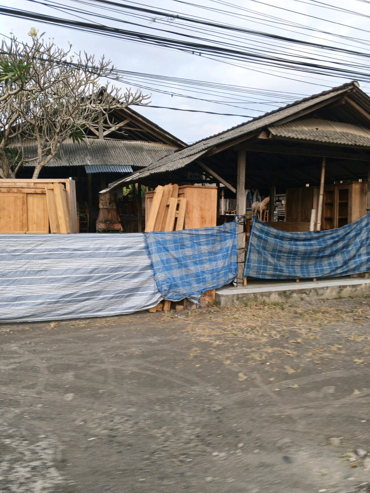 In the city of Sukawati, Indonesia, an outdoor furniture workshop is pictured. The workshop is characterized by its open-air design with a corrugated metal roof supported by wooden beams. In the foreground, two large tarps are stretched across the front, one with blue and white stripes and the other with a blue plaid pattern, obscuring the view of the workshop's interior. Behind the tarps, wooden furniture pieces are visible, including tall cabinet-like structures and stacked lumber. A bare-branched tree with sparse green leaves stands to the left of the workshop. The ground is a mixture of dark dirt and scattered dry grass. Overhead, a dense network of electrical wires crisscrosses the sky, indicative of urban infrastructure.