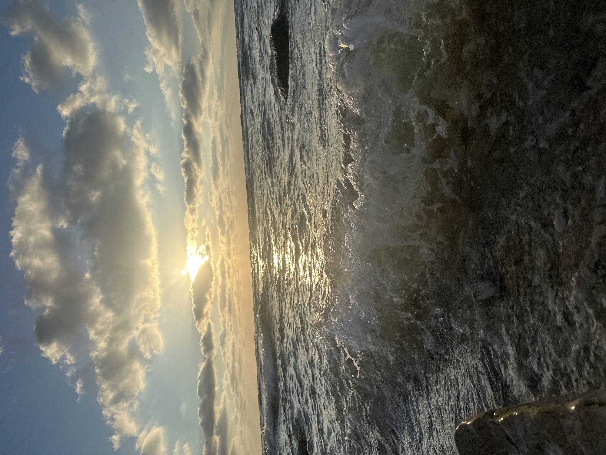This is a vertical, eye-level, close-up photograph of the sea and sky at sunset in Haifa, Israel. The sun is setting behind a cloud bank in the upper third of the image, casting a warm golden glow across the sky and water. The sea is choppy, with small waves breaking and white foam splashing near the shore. Rocks are visible at the bottom left and in the middle right of the frame. The sky is a mix of blue and white clouds, illuminated by the setting sun. There are no people or animals in the image.