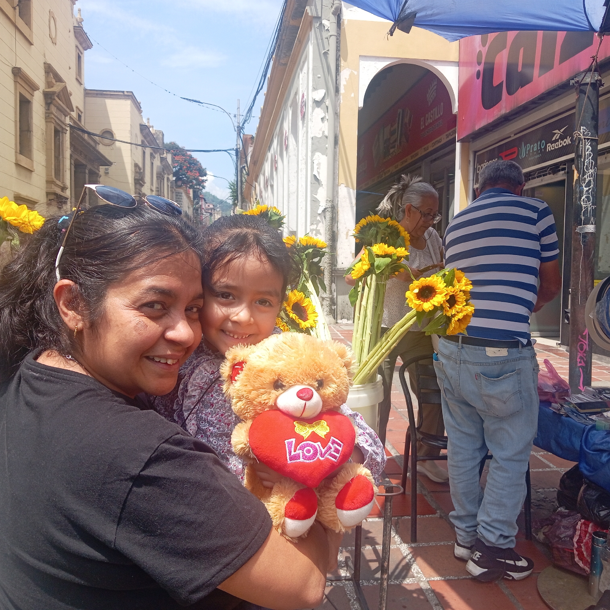 The image captures a sunny outdoor scene in a city, potentially Unknown, Unknown, featuring a woman and a girl smiling at the camera. The woman, positioned in the foreground, has dark hair and wears a black t-shirt. She is holding the girl, who is holding a teddy bear with a red heart that has the word "Love" on it. Behind them are sunflowers, and further back, an elderly woman is seen arranging more sunflowers. A man in a blue and white striped shirt stands nearby. The background includes buildings with architectural details, a blue awning, and storefront signs, including one that reads "EL CASTILLO" and another with the word "CALI". The street is paved with brick.