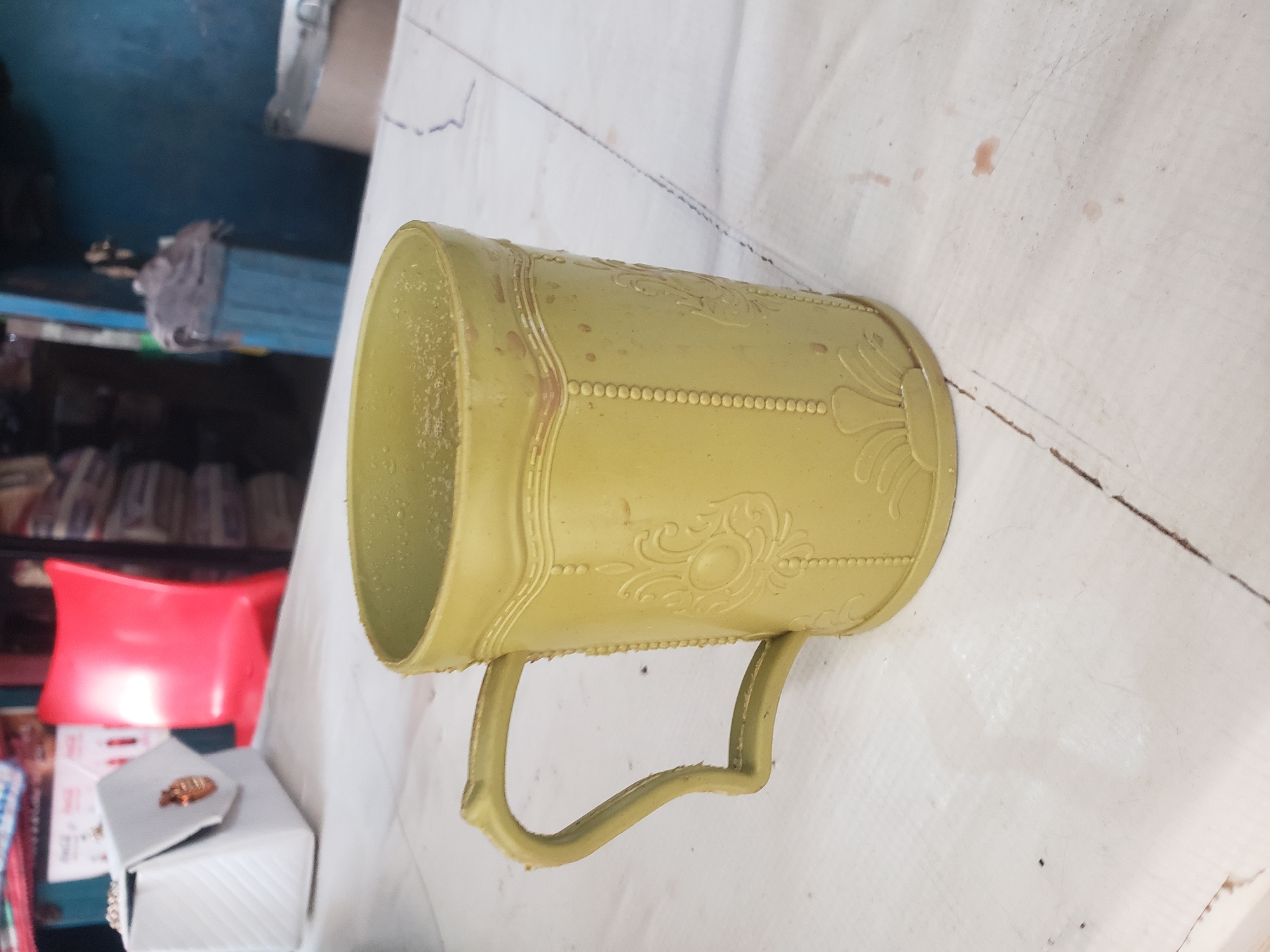 The image shows a close-up, slightly angled view of a light green plastic mug. The mug is oriented horizontally, with its handle facing downwards. The mug has a textured surface with decorative raised patterns, including a beaded border running horizontally and floral or scroll-like designs within panels. The interior of the mug is visible and appears to contain some residue.
In the background, a red object, possibly a chair or cushion, is visible. To the left of the red object, there are shelves with various items, including what appear to be books or packaged goods. The overall setting seems to be indoors, possibly a room with cluttered storage or a workshop. The lighting is diffused, suggesting an indoor environment. There is no clear indication of the time of day or weather conditions. No people are visible in the image. No discernible text is present on the mug or in the immediate surroundings. The surface on which the mug rests is white and appears to be a textured cloth or tarp, with some dark stains and scuff marks.