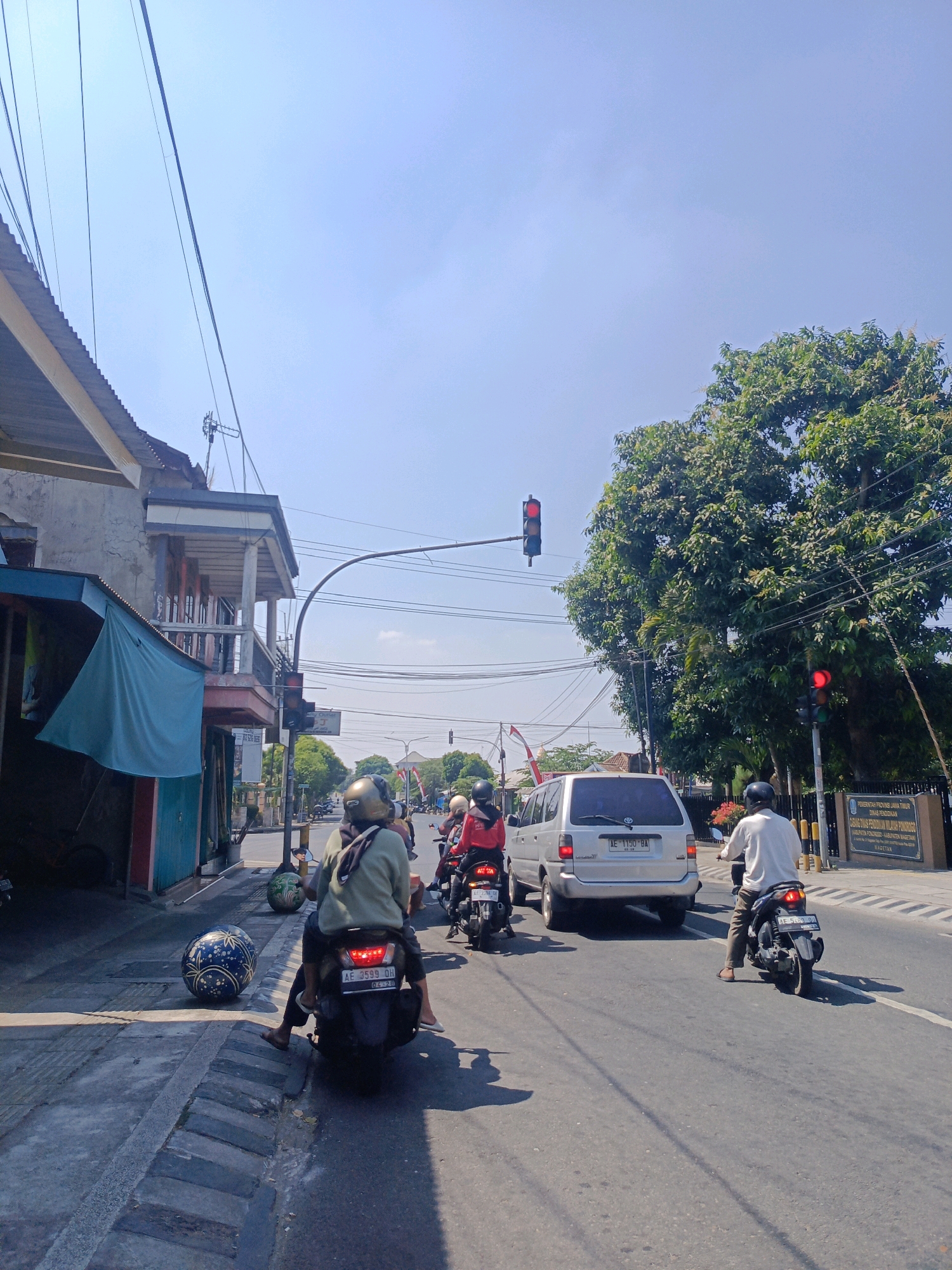 The image depicts a street scene in Sukowinangun, Indonesia, on a bright, sunny day. In the foreground, several motorcycles are stopped at a traffic light, their riders facing forward. On the left, a motorcycle with two occupants is prominent, the passenger in the back is wearing a light green jacket and a gold helmet. Next to the sidewalk on the left, a large, decorative ball with a blue and gold pattern rests on the ground. The street is paved and appears to be a main thoroughfare, with several lanes of traffic visible.
A silver van is stopped in traffic, and behind it, more motorcycles can be seen. On the right side of the road, another traffic light is visible, also showing red. A large tree with dense green foliage is situated behind the traffic light and the van.
Buildings line the left side of the street, some appearing older with weathered facades and balconies. A blue tarp is draped over a section of one building. Overhead, numerous power lines crisscross the sky, supported by poles. A sign with Indonesian text is visible on a building on the right side of the street, indicating it is likely a public institution. The overall impression is of a typical day in a bustling Indonesian city, with traffic, pedestrian activity on the sidewalk, and everyday urban infrastructure.