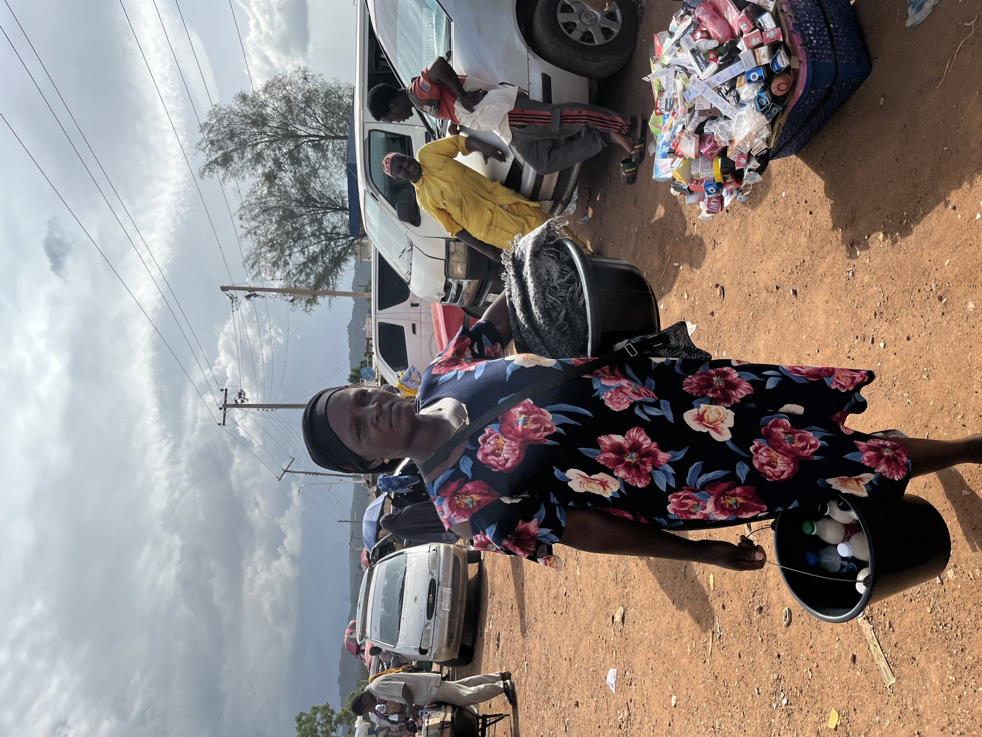 A woman wearing a black headwrap and a floral dress stands in the foreground, holding a black bucket filled with bottles of liquid. To her right, a blue basket overflows with various packaged goods. Behind her, several vehicles, including vans and cars, are parked on a dirt road. A man in a yellow garment leans against a car, talking to another person. In the background, more vehicles, tents, and people are visible. The scene appears to be a market or roadside trading area in Rafin Par, Nigeria, with a cloudy sky overhead.