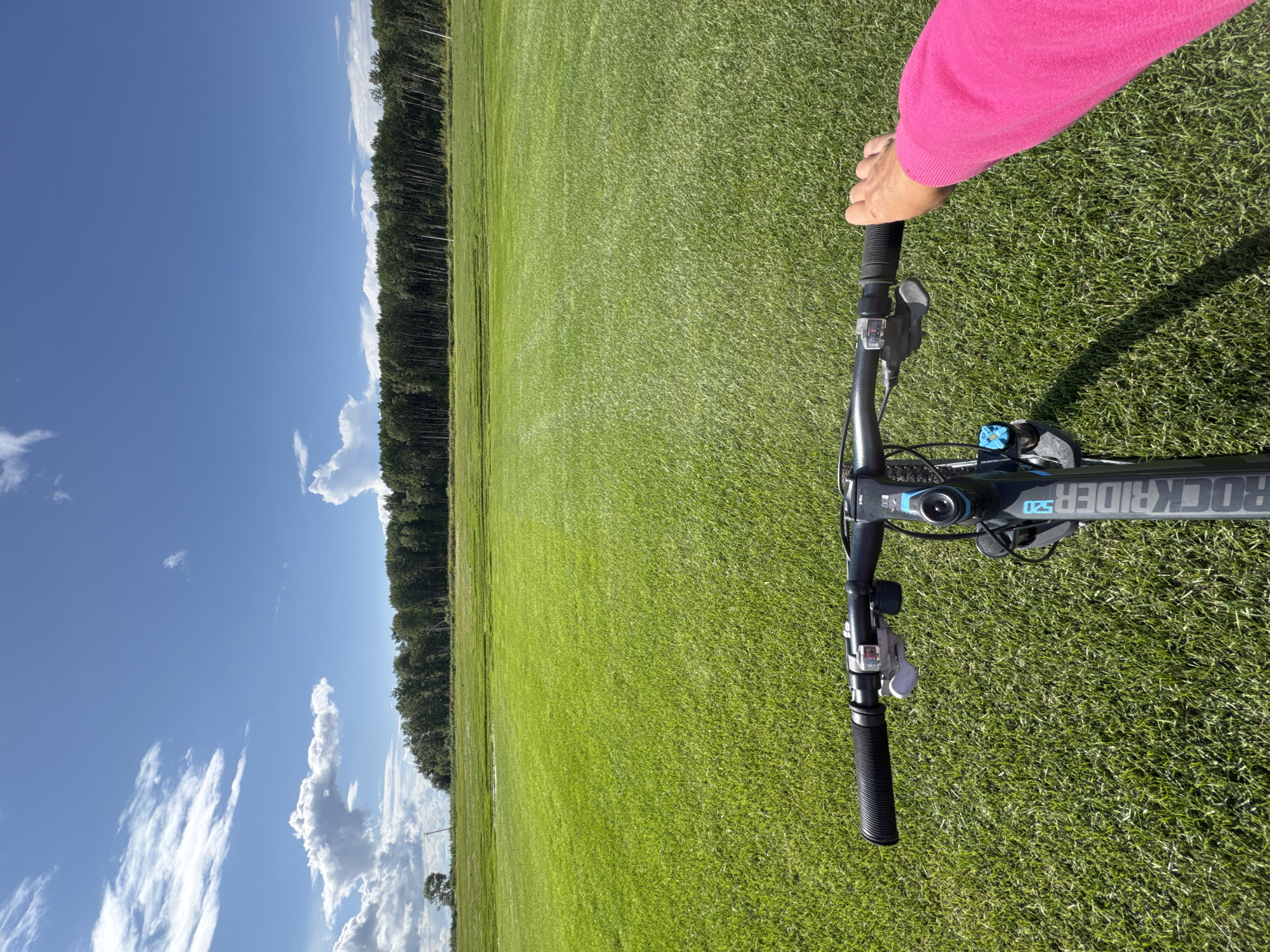 A first-person perspective shot shows a person on a bicycle on a vast, bright green field. The bicycle's handlebars are visible in the foreground, with the rider's right hand in a pink sleeve gripping the right handlebar. The field extends into the distance, meeting a dense line of trees against a clear blue sky dotted with white clouds. The scene appears to be in Innolovo, Russia, given the context of the city provided for the image.