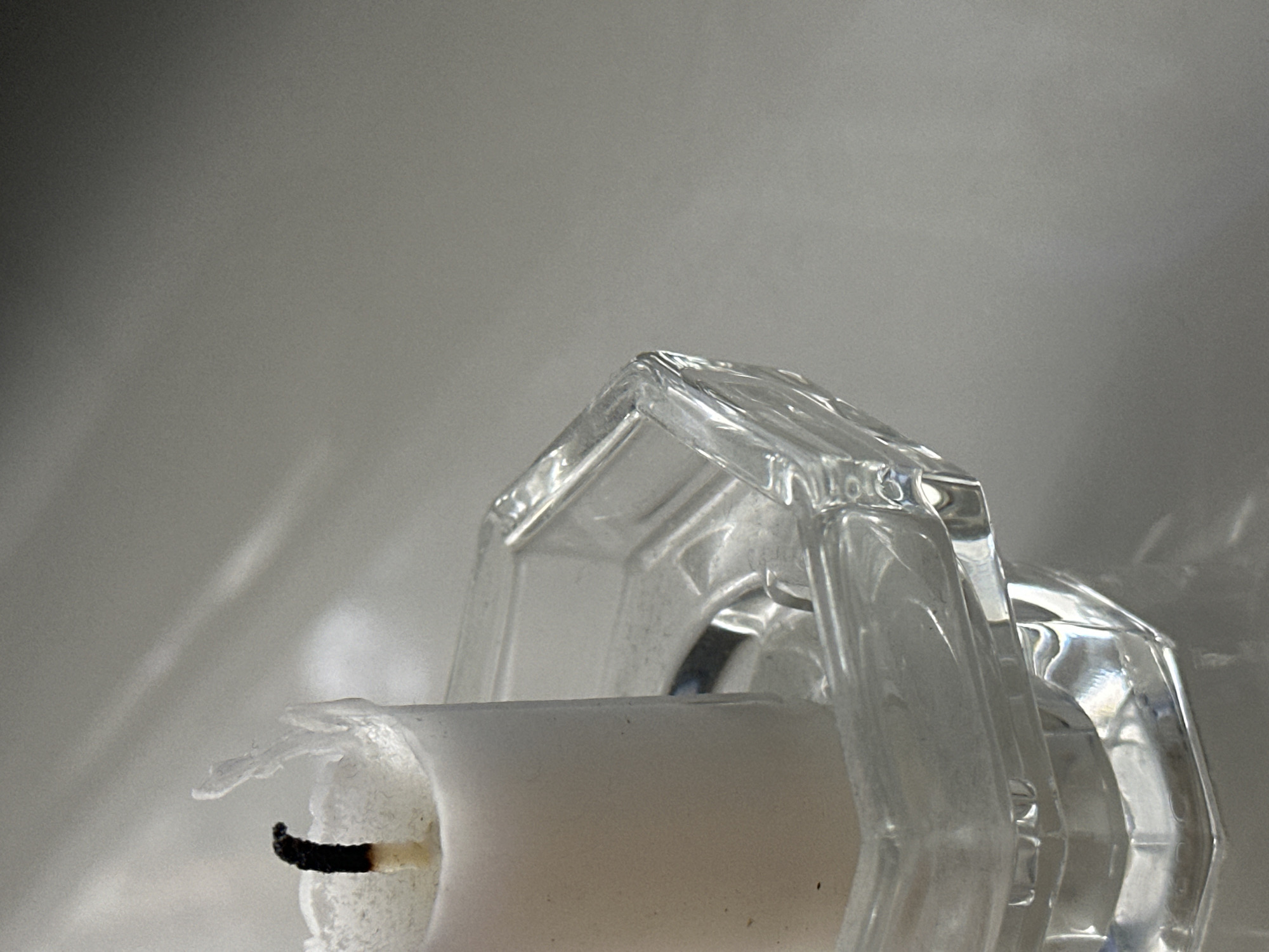 The image shows a close-up view of a partially burned white candle in a clear glass candle holder. The candle holder is octagonal with faceted sides. The wick of the candle is black and appears to be the remnant of a flame. The top edge of the candle is uneven and has melted wax drips. The background is a neutral, light gray surface with soft shadows and highlights, suggesting indoor lighting. The scene is stationary and does not depict any human presence or activity. The image does not provide any information about its location in Nevinnomyssk, Russia.