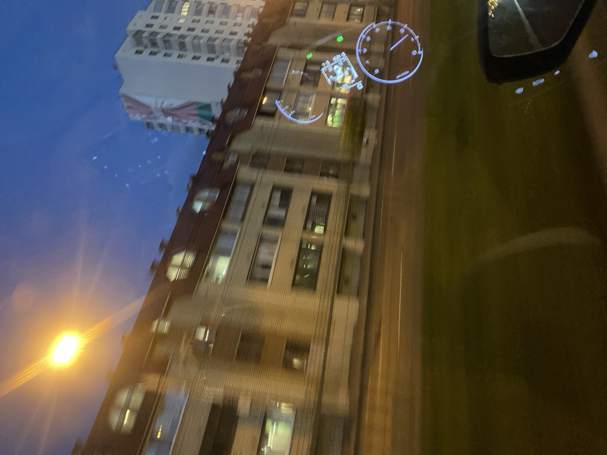 The image depicts a night-time view from within a vehicle in the city of Minsk, Belarus. The scene is characterized by motion blur, suggesting the vehicle is in transit.
Prominently featured is a multi-story apartment building with a distinctive red-tiled roof. The building's facade is a pale beige color, and it has numerous windows, some of which are illuminated, indicating occupancy. The architectural style appears to be typical of Soviet-era construction. To the right of this building, a taller, modern apartment building is visible, featuring a large banner with the colors of the Belarusian flag. The sky above is a deep blue, transitioning to dark at the top, with a bright street lamp casting a strong yellow light and creating lens flares that streak across the image.
In the foreground, a stretch of asphalt road is visible, marked with white lines. To the left of the road is a strip of green grass, indicating a roadside verge. The image also captures a reflection or overlay of a digital heads-up display (HUD) projected onto the windshield. This HUD includes circular elements resembling speedometer or dashboard gauges, with illuminated lines and markers in white and blue. There are also small green dot indicators, possibly related to navigation or vehicle systems. A portion of the vehicle's side mirror is visible in the bottom right corner of the frame, reflecting streetlights.
No people are clearly discernible in the image due to the motion blur and the night-time setting. The overall impression is one of urban travel at dusk or night, with elements of both traditional and modern architecture characteristic of Minsk. The specific location within Minsk is not identifiable from visual cues alone, beyond the general urban context and the presence of the Belarusian flag.