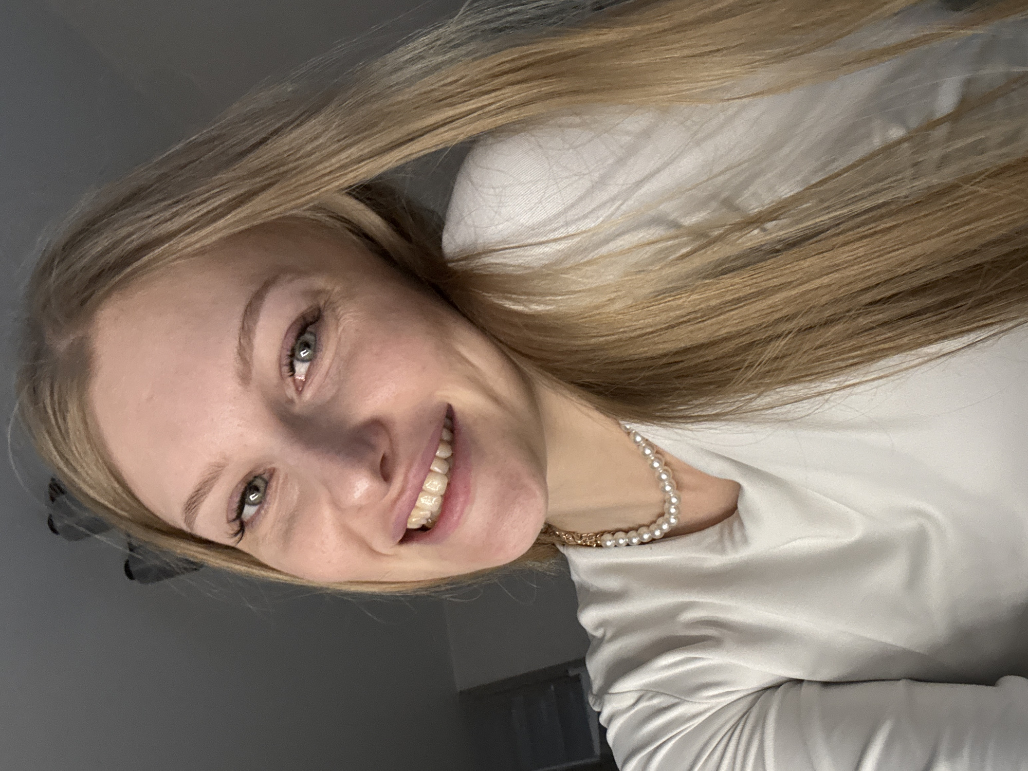 A close-up, angled portrait captures a young woman with long, straight, light blonde hair. She is smiling broadly and looking directly at the camera with light-colored, possibly green or hazel eyes, conveying a happy and friendly demeanor. She wears a light, smooth-textured long-sleeved top, appearing white or very light gray. Around her neck is a necklace featuring spherical pearls interspersed with a small section of gold chain. A clear dental appliance, possibly a retainer or aligner, is visible on her upper teeth. The background is dark and out of focus, suggesting an indoor setting. There is no visible text in the image. The location context provided is Perm, Russia.