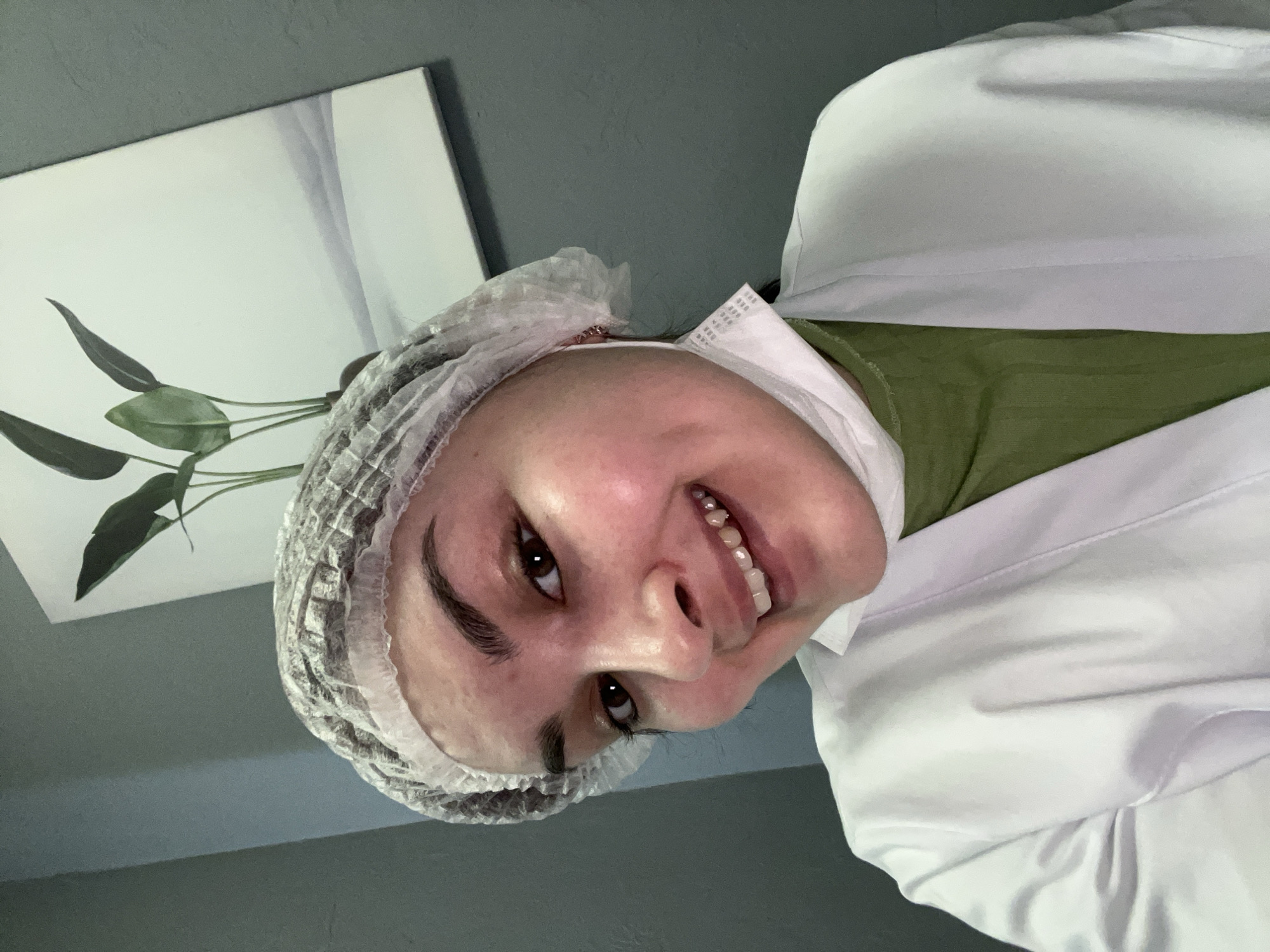 A woman wearing a white medical lab coat and hairnet is looking at the camera. She is also wearing a green shirt and what appears to be a disposable medical mask pulled down. A painting with a plant on it is visible on the wall behind her. The location is unknown, and there is no evidence to suggest that it is in Neftekamsk, Russia.