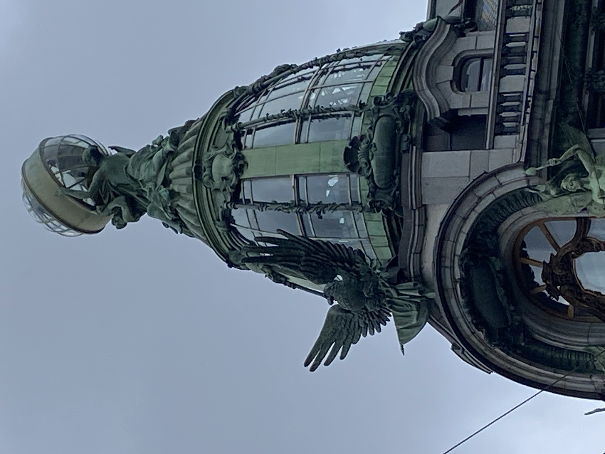 The image shows the Singer House, also known as the Dom Knigi (House of Books), located on Nevsky Prospekt in Saint Petersburg, Russia. The photograph captures a close-up view of the building's iconic dome and rooftop architectural elements. Prominently featured is a large, green bronze eagle with outstretched wings, perched on a decorative architectural element. Above the eagle, the glass dome of the Singer House is visible, topped by a sculpted globe held aloft by figures. The building's facade, characterized by intricate detailing and ornate sculptures, is also evident. The sky in the background is overcast and gray, suggesting a cloudy day. The image is taken from a low angle, emphasizing the grandeur and height of the structure.