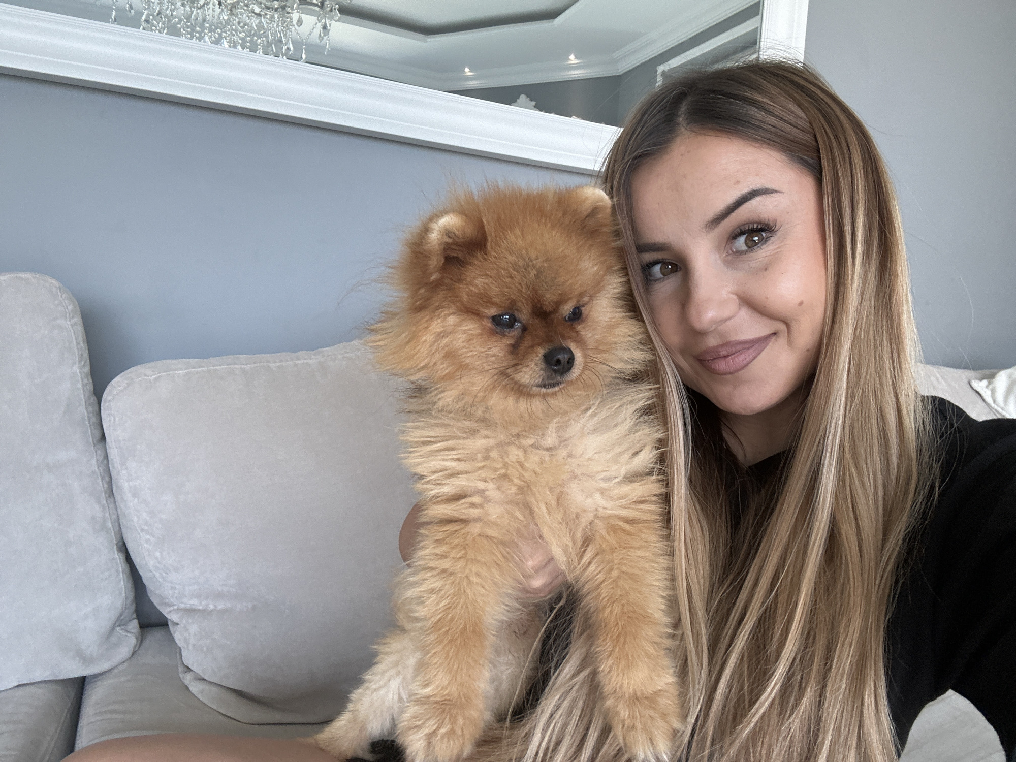 A young woman with long, blonde-brown hair and light makeup is holding a small, fluffy Pomeranian dog. She is smiling and looking at the camera. The dog, which is a golden-brown color, is nestled against her shoulder. They are seated on a light-colored couch, which is part of a living room. In the background, a wall painted a muted blue is visible, and above that, a white framed mirror reflects a room with a chandelier. The overall setting appears to be a well-decorated indoor space, likely a home. There is no specific indication of the city Asenovgrad, Bulgaria in the image.