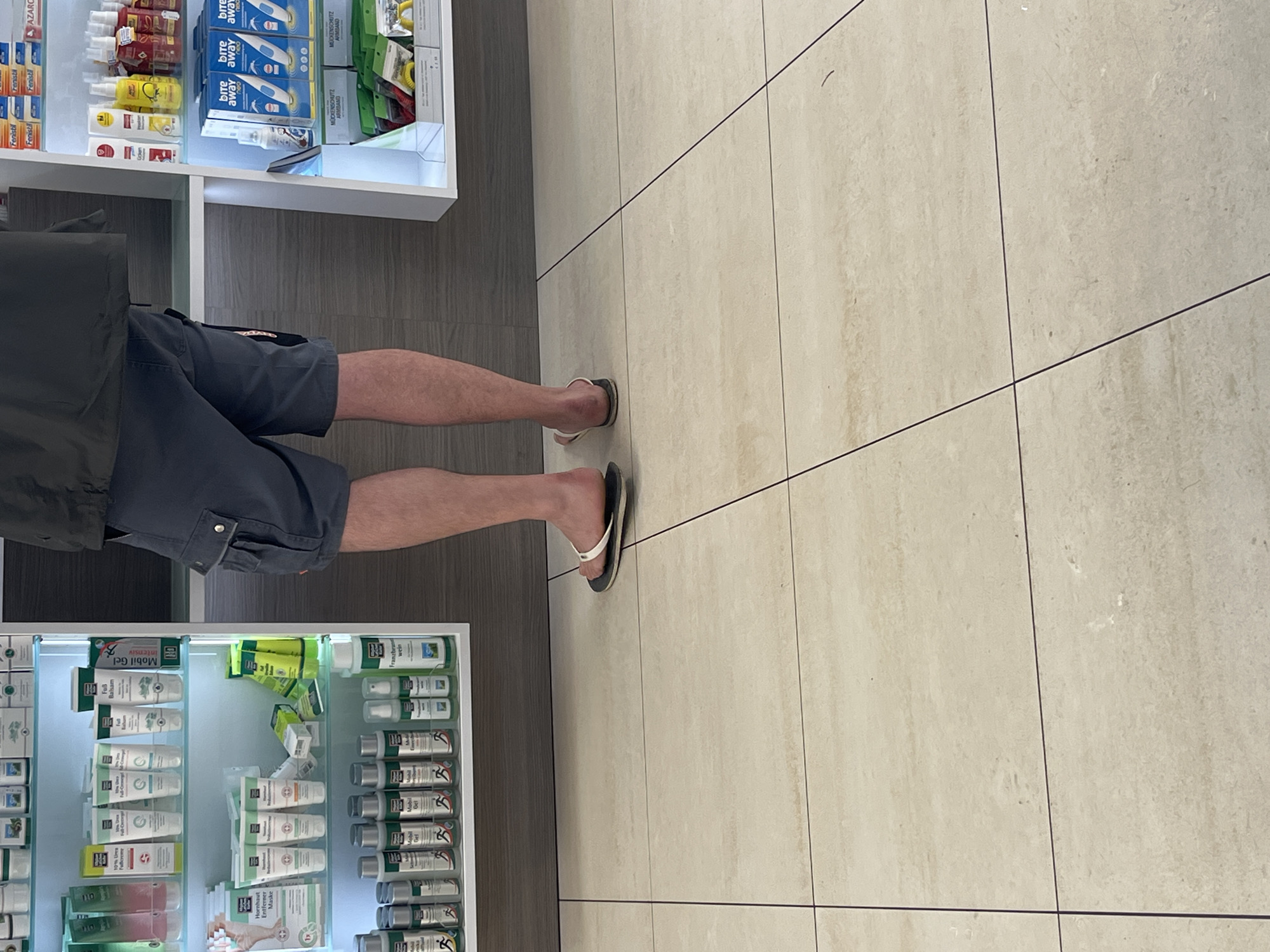 The image shows a person standing in what appears to be a pharmacy or drugstore. The person is wearing gray shorts, a dark green jacket, and flip-flops. The person is standing in front of two shelves filled with various products, including medicines and personal care items. One shelf contains products such as "Mobil Gel", "Franzbranntwein", and "Fenistil". Another shelf has products with the brand "bite away" and "MÜCKENSCHUTZ ARMBAND". The floor is tiled with light-colored tiles. Based on the presence of German brand names like "Franzbranntwein", "Fenistil", "Mobil Gel" and "MÜCKENSCHUTZ ARMBAND", and the possible brands being sold, it is likely a pharmacy in Germany. It is impossible to specify the exact location or city without more context, but based on the brands and available information, it is likely in a German-speaking region. There's no information to confirm it's in Saarbrücken.