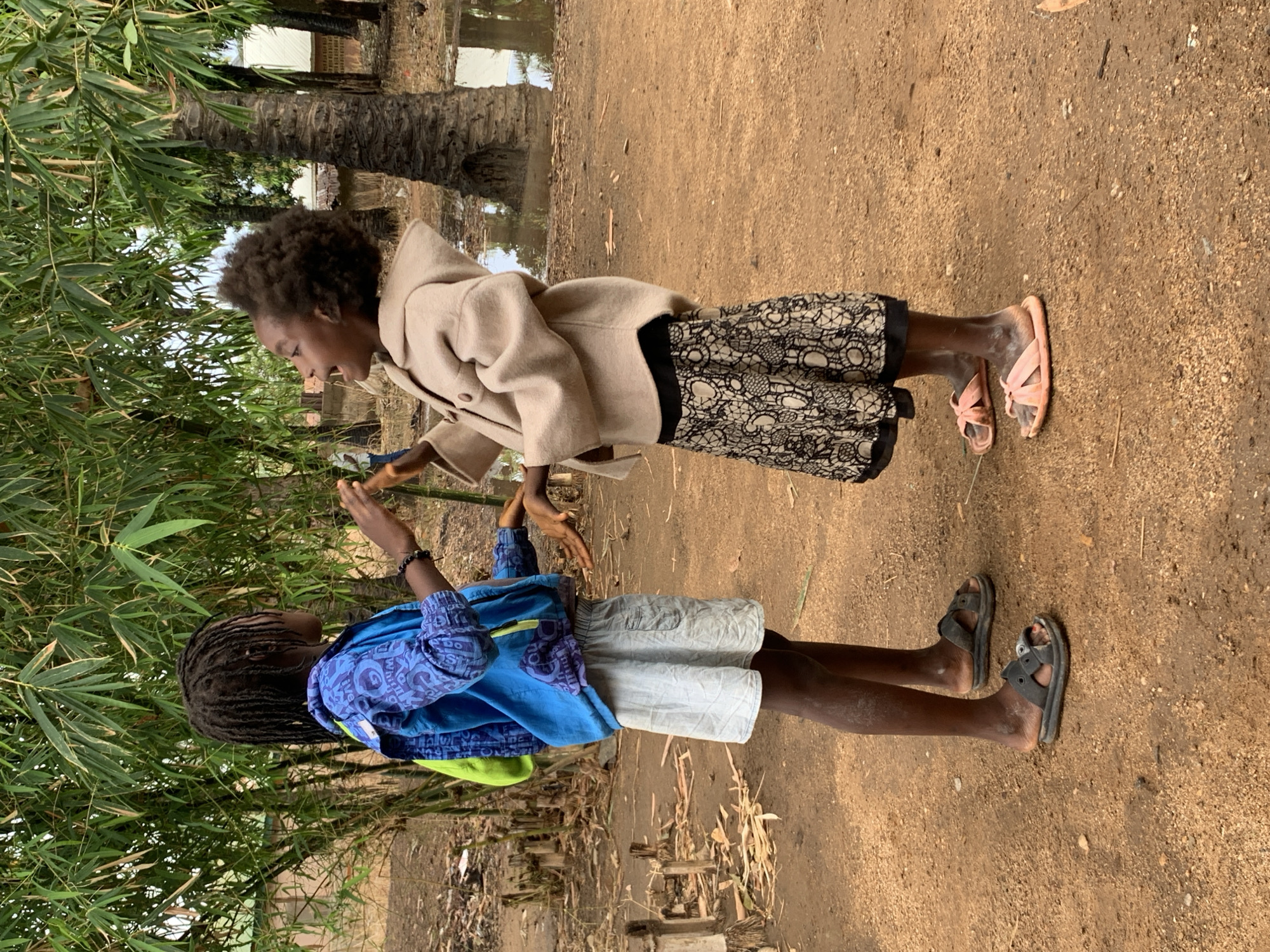 Two children stand on a dirt ground near bamboo shoots in Akwanga, Nigeria. One child, with braided hair, wears a blue patterned shirt, light shorts, a green backpack, black sandals, and a bracelet. The other child, with afro-textured hair, wears a tan coat, a black and lace patterned skirt, and pink sandals. They are facing each other, with their hands in the air, seemingly interacting. A tree stands in the background near a puddle.