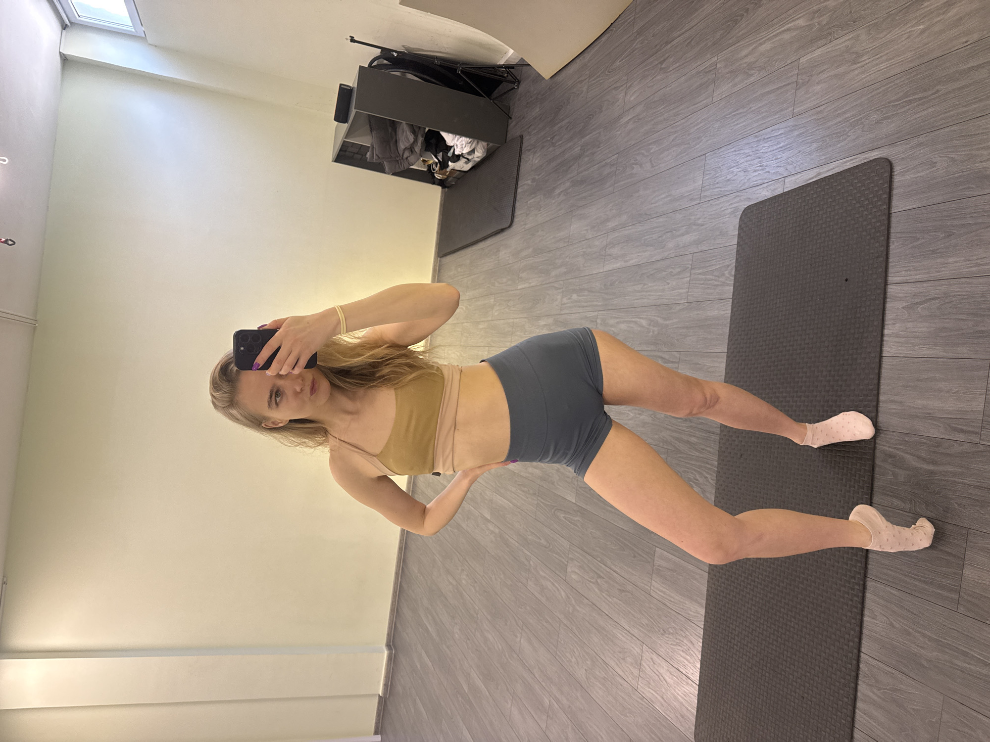 A full-length shot shows a blonde woman standing in a room, likely an indoor workout space, holding a phone at eye level to take a selfie. She is wearing a light brown sports bra and blue-grey shorts with socks. She is standing on a grey yoga mat on a floor that is grey with a wood grain pattern. Behind her is a light-colored wall with a closet visible on the far wall.