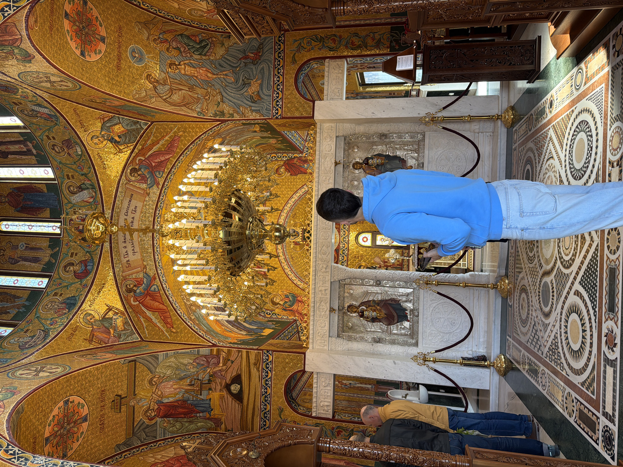 The image shows the interior of the Cathedral Church of the Assumption in Plovdiv, Bulgaria. The scene is dominated by the ornate, mosaic-covered ceiling, featuring religious iconography. A large chandelier hangs from the center of the ceiling. On the right side of the frame, a man in a blue sweatshirt and jeans stands at a barrier, seemingly taking a picture with his phone. In front of him, there is a small marble structure with an icon. In the foreground, another person is seen praying or bowing towards the icon. The floor is tiled with a patterned design. The overall environment appears to be richly decorated and religiously significant.