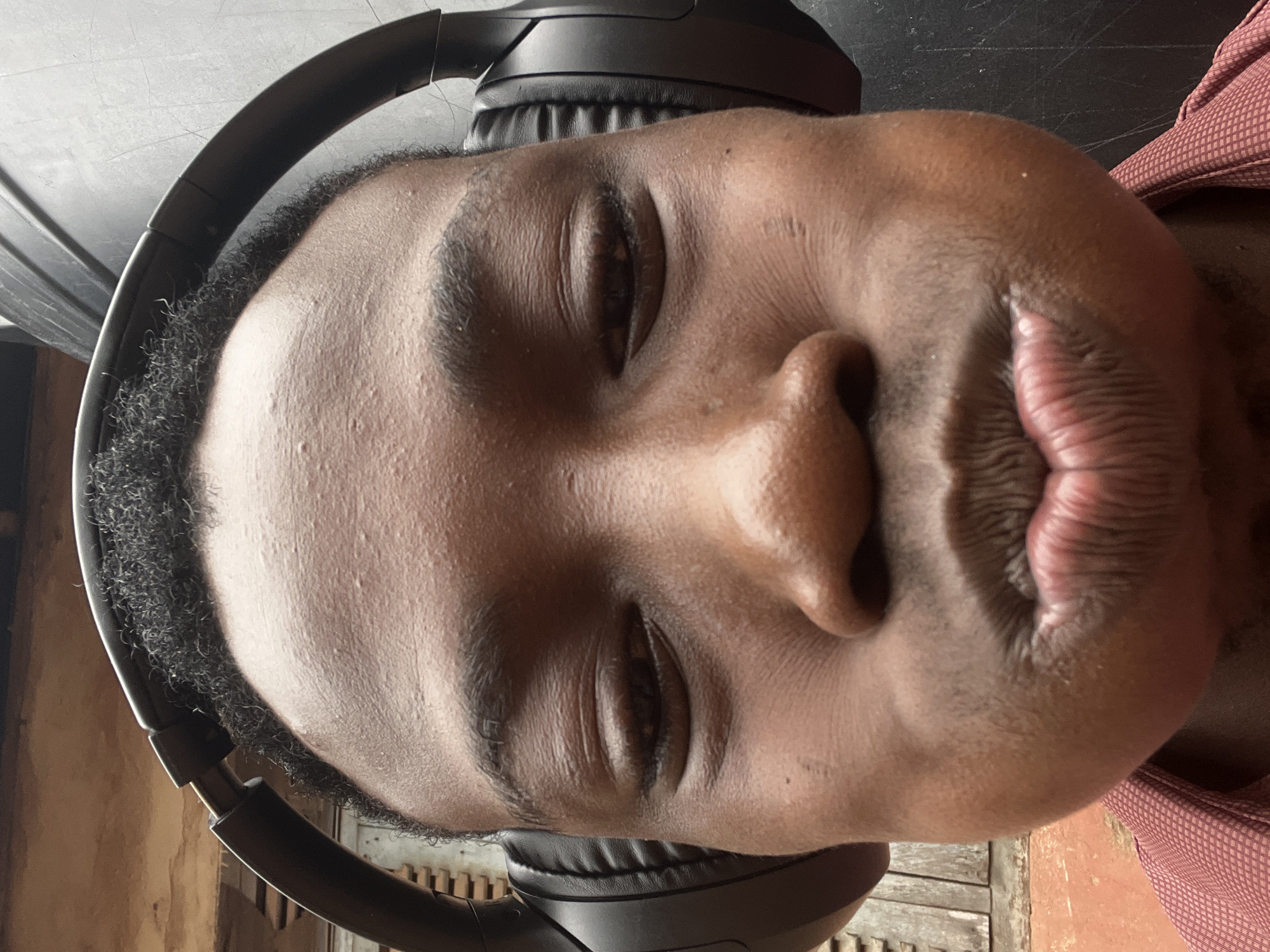 A close-up, selfie-style photo shows a young Black male wearing large black over-ear headphones. The subject's head is tilted slightly, and his eyes are closed, with his cheeks puffed out and his lips pursed in a pout. The background is blurred but appears to be outdoors, with elements that suggest an urban environment in Onitsha, Nigeria, possibly a wall or building to the left and a dark, textured surface to the right. The subject is wearing a reddish-brown collared shirt.