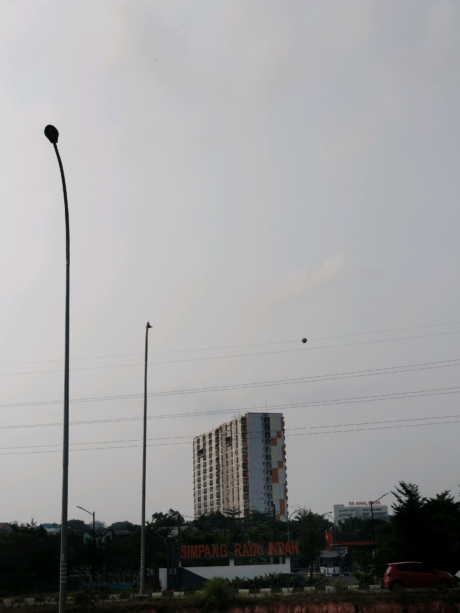 The image depicts an overcast sky over a cityscape in Batam, Indonesia, likely near a suburban or developing area. In the foreground, two tall, dark streetlights stand prominently on the left and center-right of the frame. The left streetlight is a solitary, slender pole with a rounded top, while the right one is slightly shorter and has a bird perched on its apex. Several parallel power lines stretch horizontally across the middle of the image, with a small, dark spherical object, possibly a ball, suspended on one of them.
Dominating the mid-ground is a tall, multi-story building with a light-colored facade accented with orange and white sections. Its numerous windows are visible, suggesting an apartment complex or office building. Further to the right, a smaller building with "RS AWAL BROS" is visible, indicating a hospital. In the lower portion of the frame, a large orange sign with white lettering reads "SIMPANG RAYA INDAH," identifying a location or development. Trees and other foliage form a dark line across the bottom, interspersed with some structures. A red car is parked on the right side of the image, near the sign. The overall impression is of an urban or semi-urban environment under a muted sky.