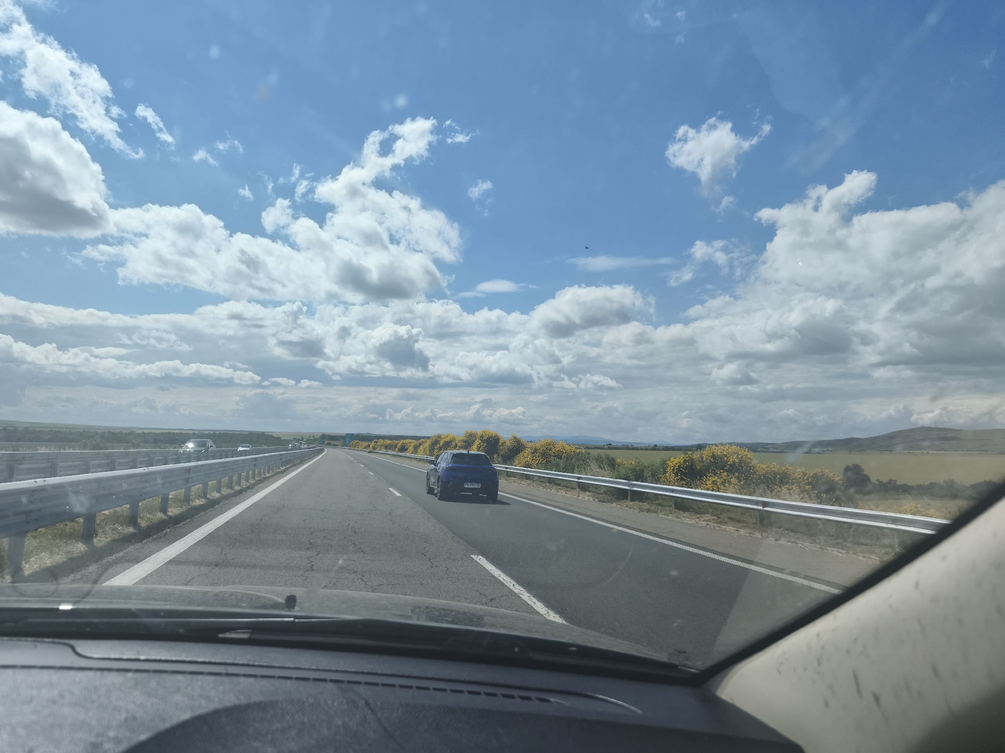 This is a forward-facing, wide shot of a highway with a blue car driving in the right lane. The road is paved and has white lane markings. The highway is bordered by a metal guardrail on the left and yellow flowering bushes on the right. In the distance, there are rolling hills and a blue sky with white clouds. The image is taken from inside a vehicle, with the dashboard visible in the foreground. The location is the highway near Kozare, Bulgaria.