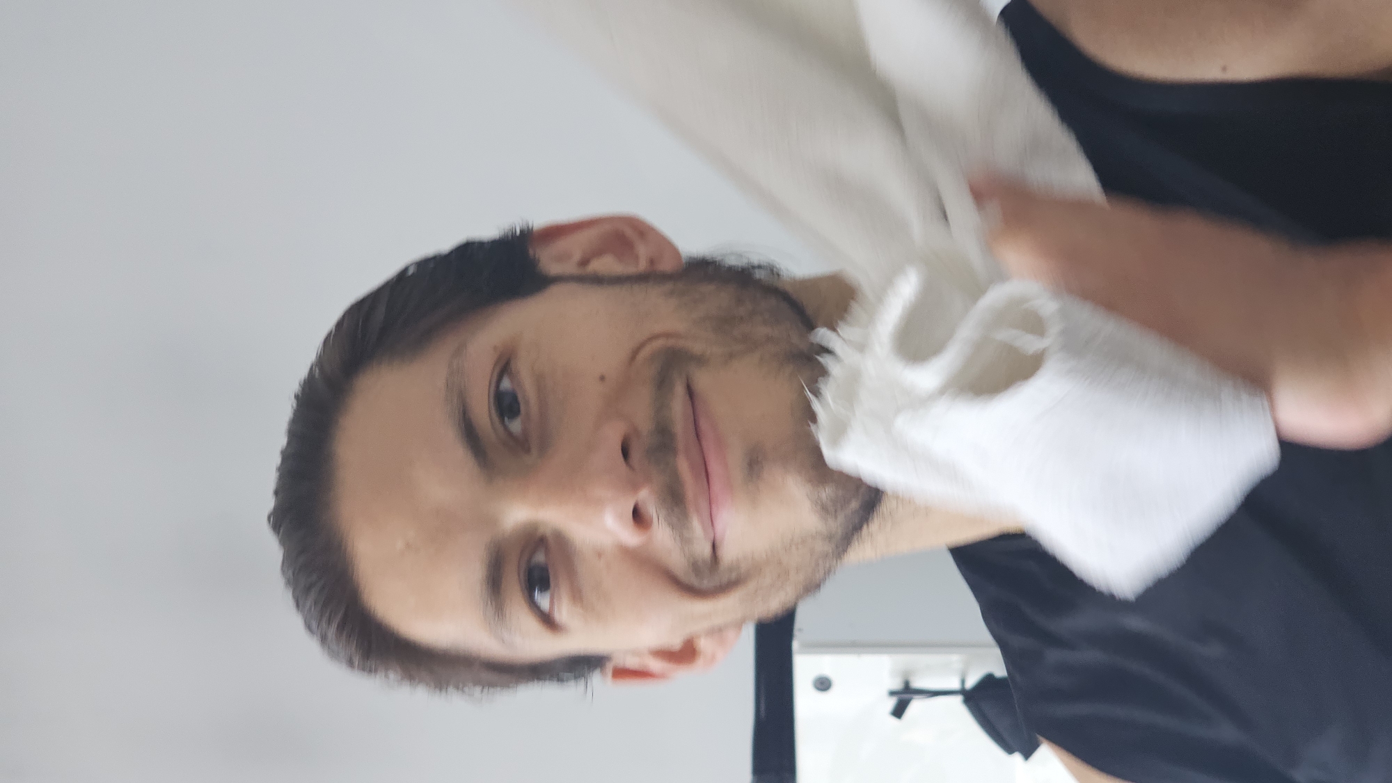 A close-up, portrait-style shot captures a man from the chest up, positioned against a plain, light-colored background. He has dark hair, styled back, and dark facial hair (stubble/beard). The man is looking directly at the camera with a slight, pleasant smile or smirk on his face.
He appears to be in the process of wiping or drying himself with a white, somewhat textured cloth or towel, which is held in his right hand and partially obscures the right side of his face and neck. He is wearing a dark, possibly black, short-sleeved top or t-shirt, visible around his shoulders and chest. In the lower right background, a glimpse of a darker, possibly black, object with a 'T'-shaped element suggests an indoor setting, perhaps a fixture on a wall or a piece of furniture.
The lighting is bright and even, consistent with an indoor environment during the daytime. The man's expression conveys a relaxed and possibly amused or friendly demeanor. There is no visible text in the image. The location context provided is Puebla, Mexico, though no specific visual cues within the image confirm this.