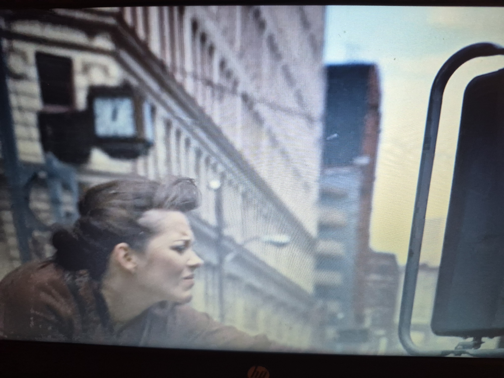 The image shows a woman with an unusual hairstyle, with her hair pulled back and shaved on the sides, wearing a brown garment. She is looking down and to her right with a concerned expression. The background appears to be a city street with tall buildings. On the left side of the image, there are two dark rectangular objects mounted on a wall, one with a glowing blue light inside. A railing runs along the side of the scene, suggesting an elevated walkway or bridge. In the foreground, to the right, is a dark object with a curved metallic frame, possibly part of a vehicle or structure. The overall scene is blurred, indicating it might be a still from a video or a photograph taken with a low-resolution camera. There is no discernible information within the image to suggest a specific location in Sandwell, United Kingdom.